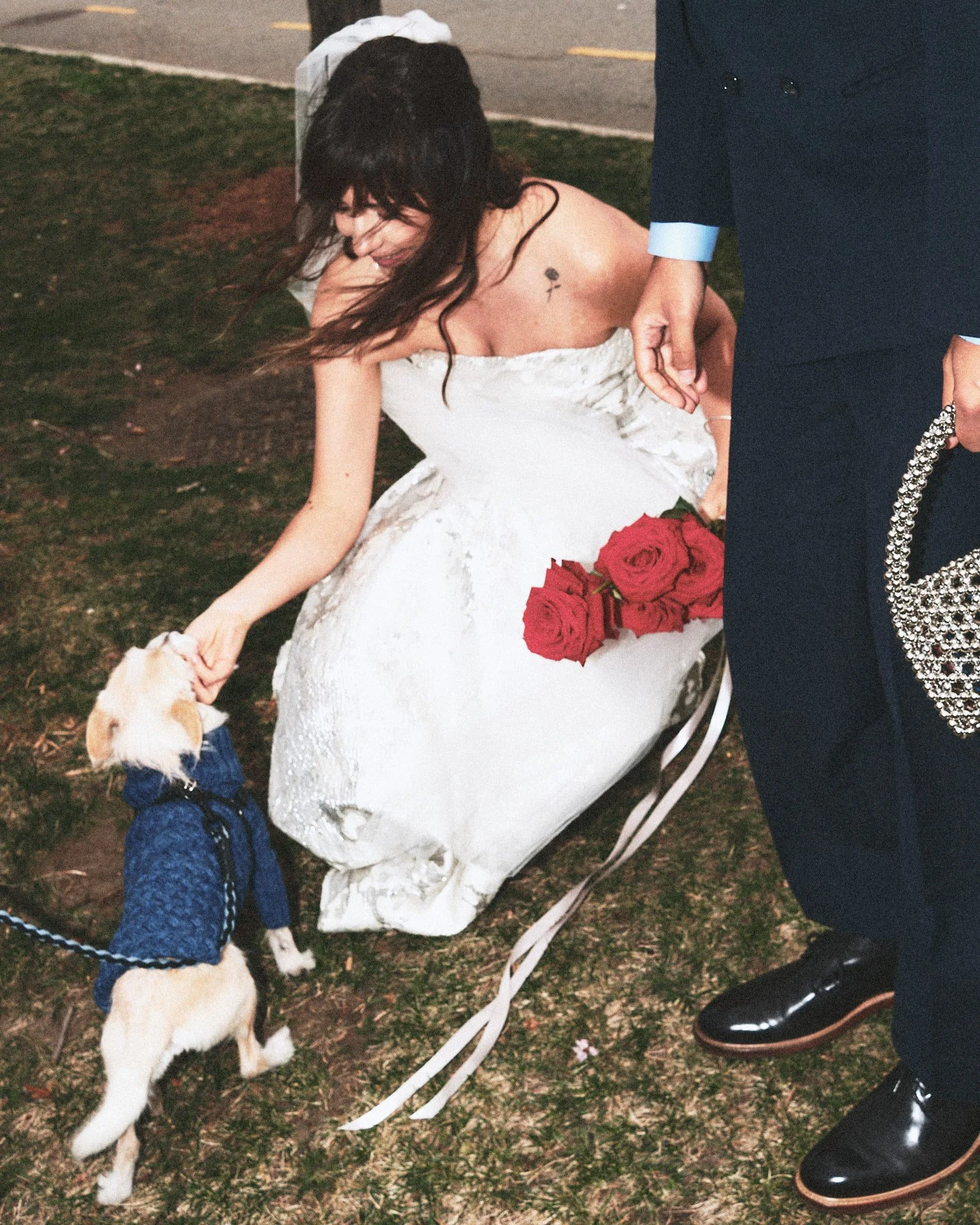 A woman in a wedding dress kneeling on the ground holding the leash of a dog, with a man standing next to her. The woman has long dark hair, a small tattoo on her upper chest, and is smiling. The dog is wearing a denim jacket and looking up at the woman. There are red roses on the ground near her. The scene appears to be outside at night.