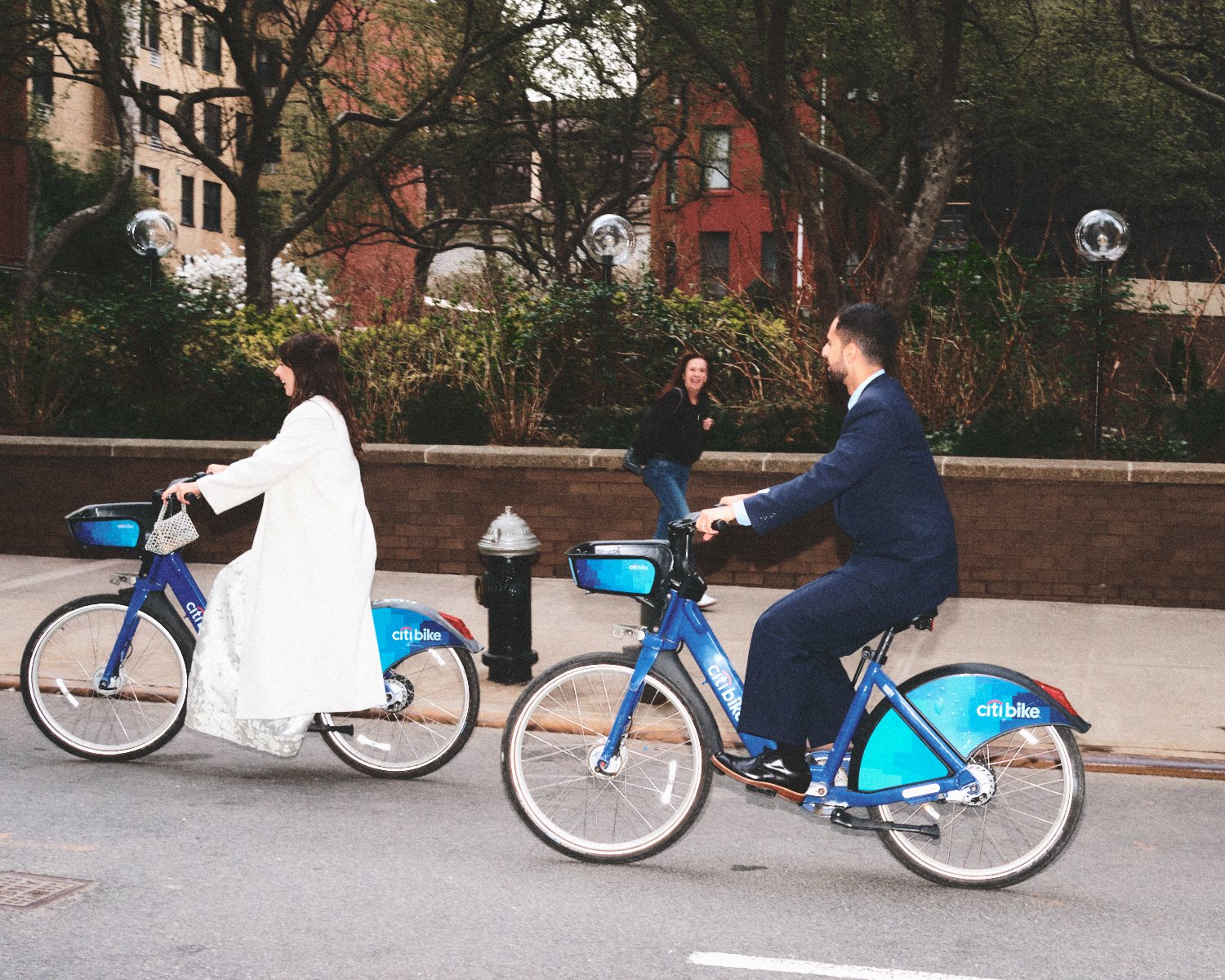 A man and woman riding blue Citi bikes, photographed by Madivan Studios, on a NYC city street with a woman walking and smiling in the background.