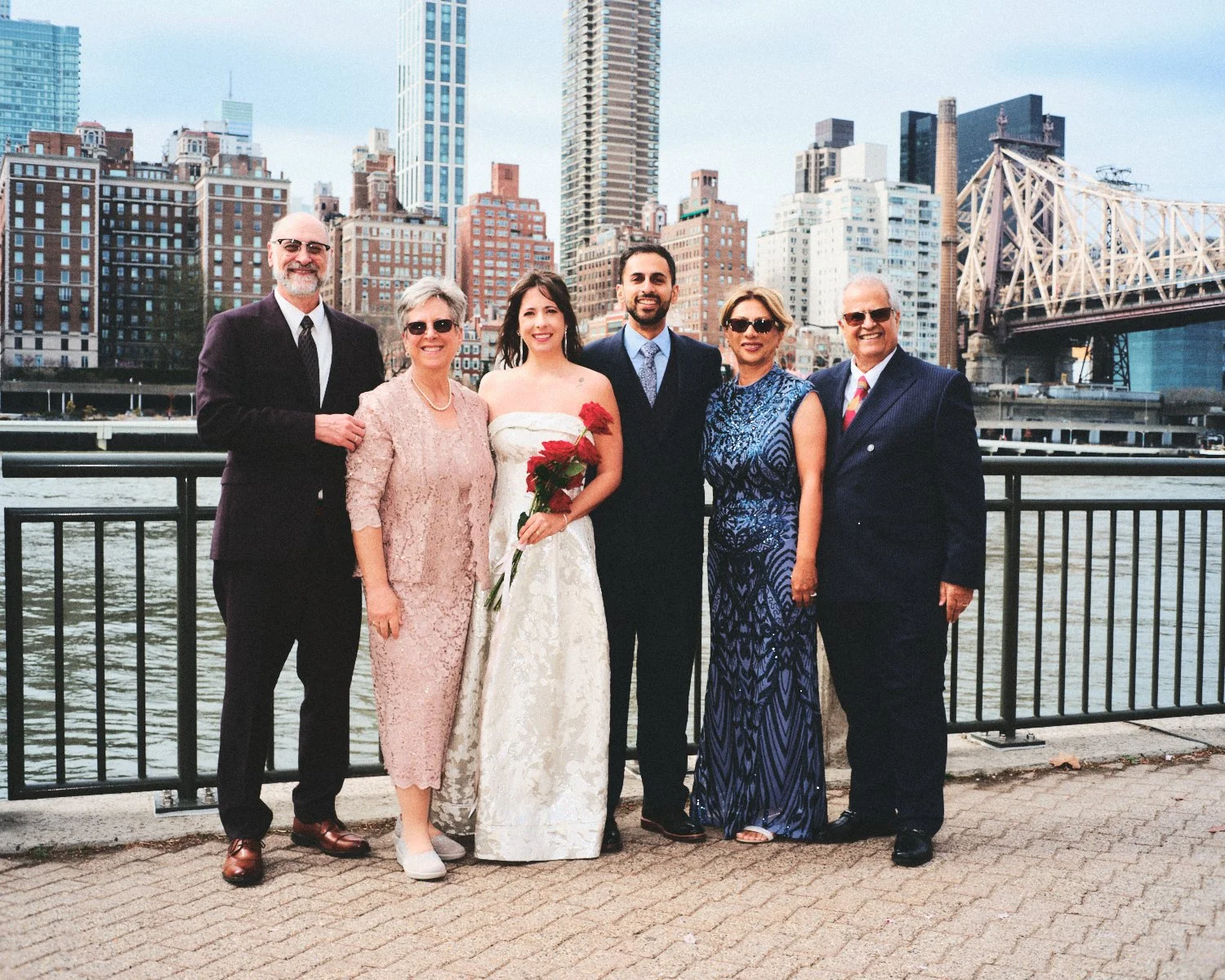 A group of six people, including a bride and groom, standing outdoors by the water with the New York City skyline and bridge in the background, during daytime photographed by Madivan Studios.