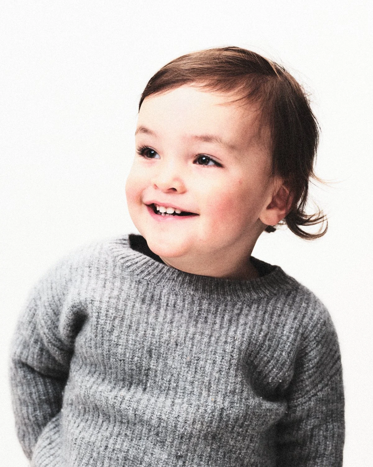 A young boy with dark hair smiling and wearing a gray knit sweater, standing against a plain white background. Photographed in monochrome by Madivan Studios in New York City.