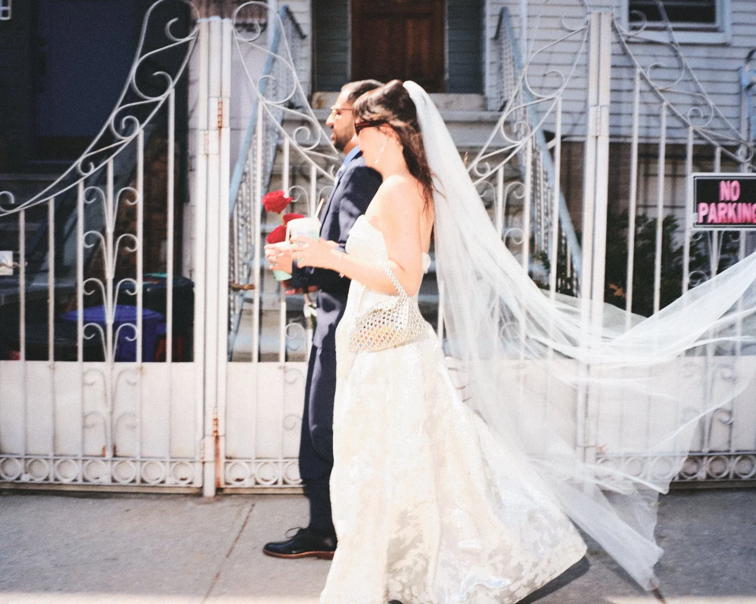 A bride and groom walking past a white wrought-iron NYC gate, with the bride in a wedding dress and veil holding a bouquet of red roses, and the groom in a suit holding a drink photographed by Madivan Studios.