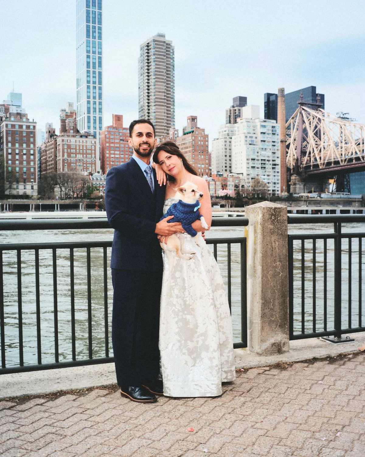 A newlywed couple in formal attire standing by a waterfront photographed by Madivan Studios, with the NYC skyline and bridge in the background, holding a small dog in a blue sweater.