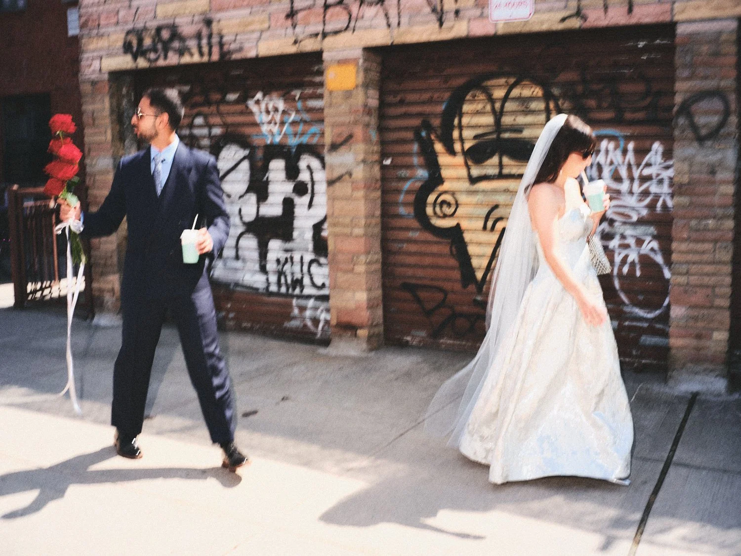 A man in a dark suit and sunglasses holding a bouquet of red roses and a drink, looking at a woman in a wedding gown with a veil, who is also carrying a drink, walking past a NYC graffiti-covered brick wall photographed by Madivan Studios.