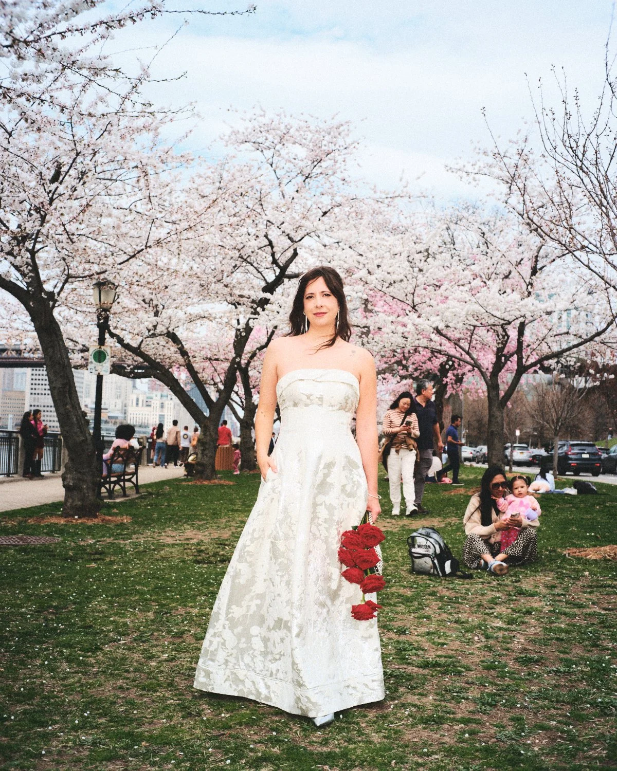 A woman in a white strapless dress, photographed by Madivan Studios, standing on a grassy area under blooming cherry blossom trees in New York City, holding a garland of red roses, with people in the background during spring.
