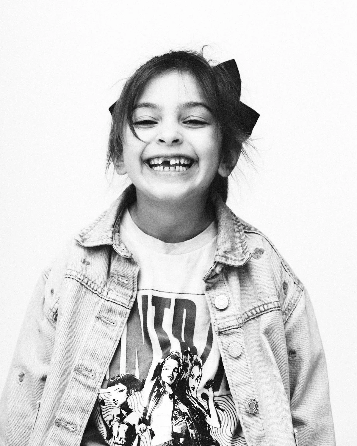 A smiling young girl with missing front teeth, wearing a graphic t-shirt and denim jacket, with a large bow in her hair, against a plain white background. Photographed in monochrome by Madivan Studios in New York City.
