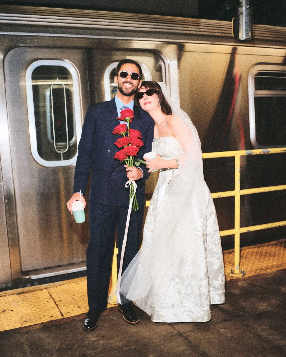 A couple dressed in wedding attire standing next to a silver train on a New York City subway platform at night, with the man holding a bouquet of red roses and the woman leaning on him, photographed by Madivan Studios.