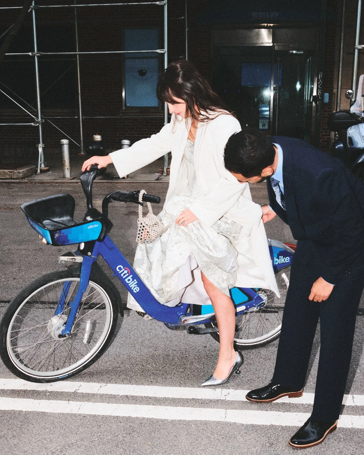 A woman in a white wedding dress and heels getting on a CitiBike in NYC with a man in a dark suit helping her. They are on a city street at night and photographed by Madivan Studios.