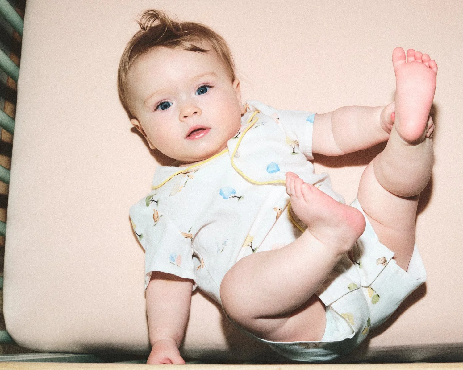 Documentary flash photography of a baby lying on a bed in a New York City apartment, wearing white pajamas with animal prints. The baby has blue eyes and brown hair, and is stretching while looking at the camera being photographed by Madivan Studios.