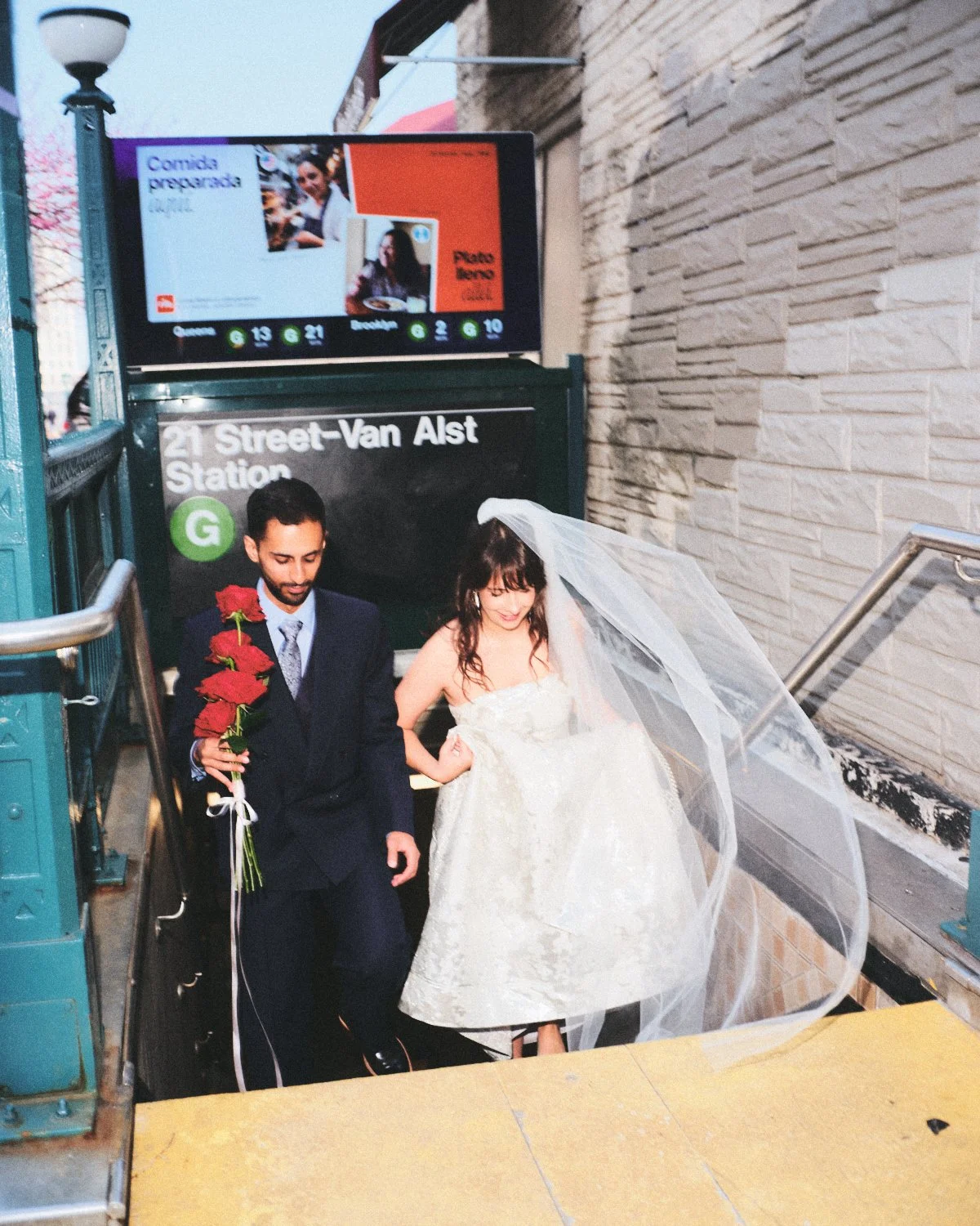 A newlywed couple photographed by Madivan Studios walking up the stairs at 21st Street-Van Alst Station in NYC, with the bride wearing a wedding gown and veil, and the groom in a suit holding a bouquet of red roses.
