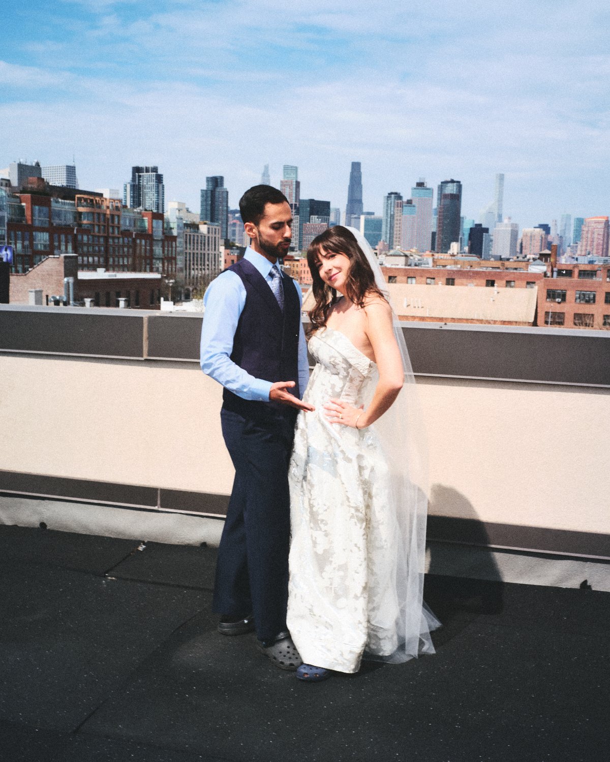 A man and a woman photographed by Madivan Studios in wedding attire standing on a rooftop with the NYC skyline in the background.