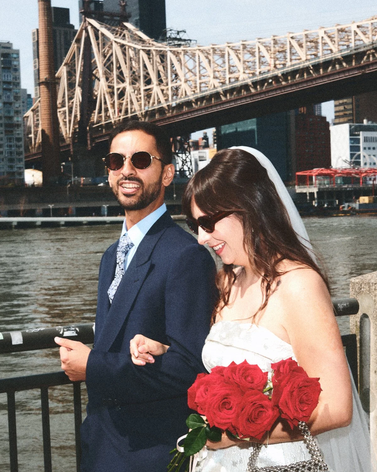 A happy couple in wedding attire photographed by Madivan Studios standing near a river with the NYC skyline and bridge in the background, holding a bouquet of red roses.