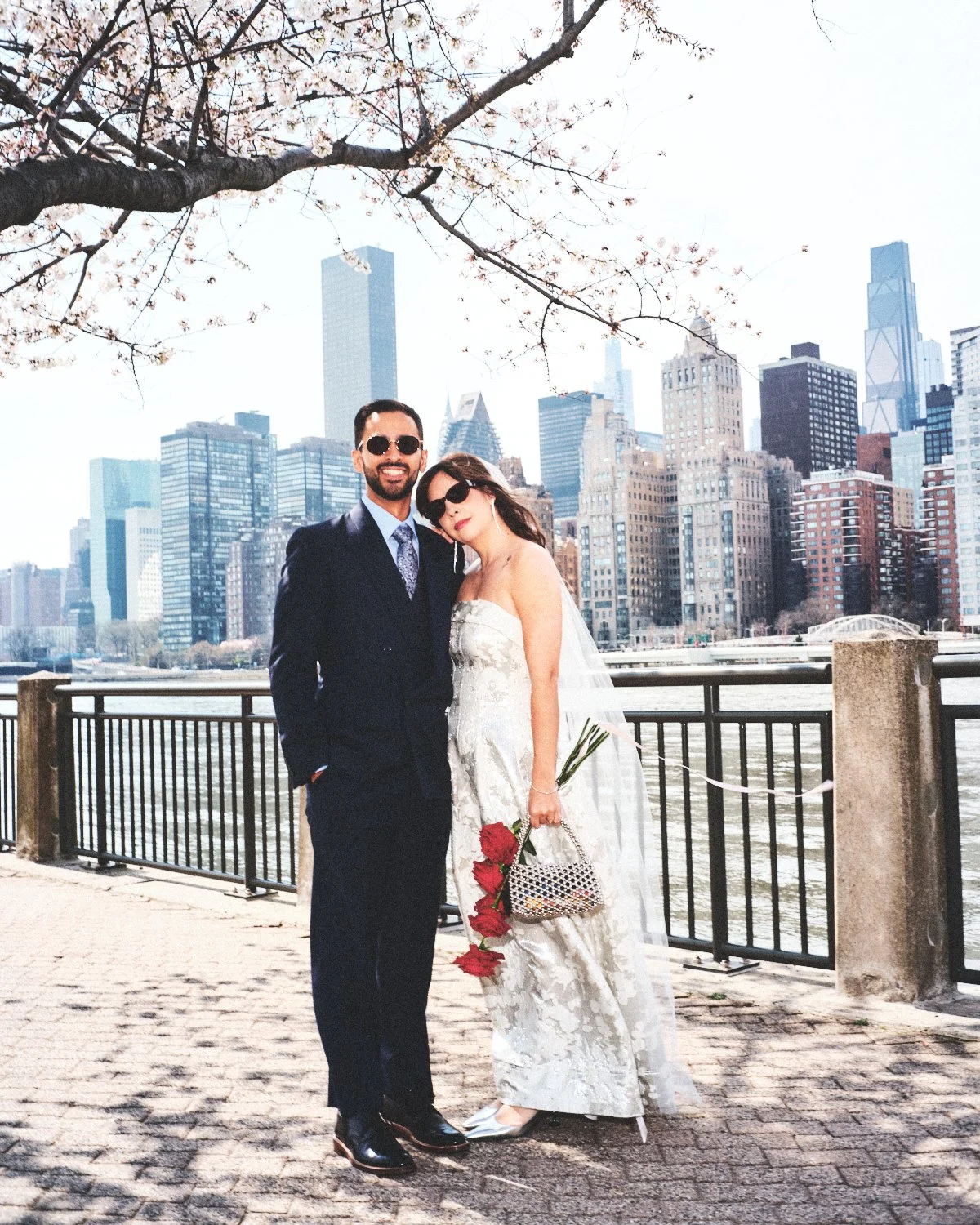 A newlywed couple dressed in formal attire photographed by Madivan Studios, standing on a sidewalk by a waterfront with the NYC skyline in the background, under a flowering tree.