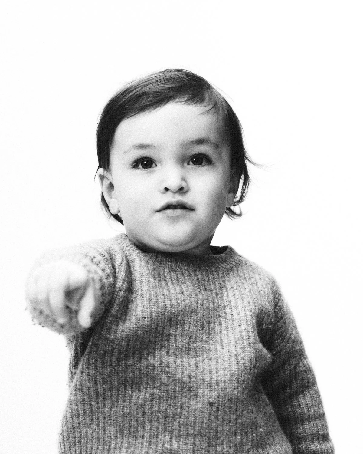 Black and white photo of a young child with short dark hair, wearing a knitted sweater, pointing directly at the camera with a neutral expression. Photographed in monochrome by Madivan Studios in New York City.