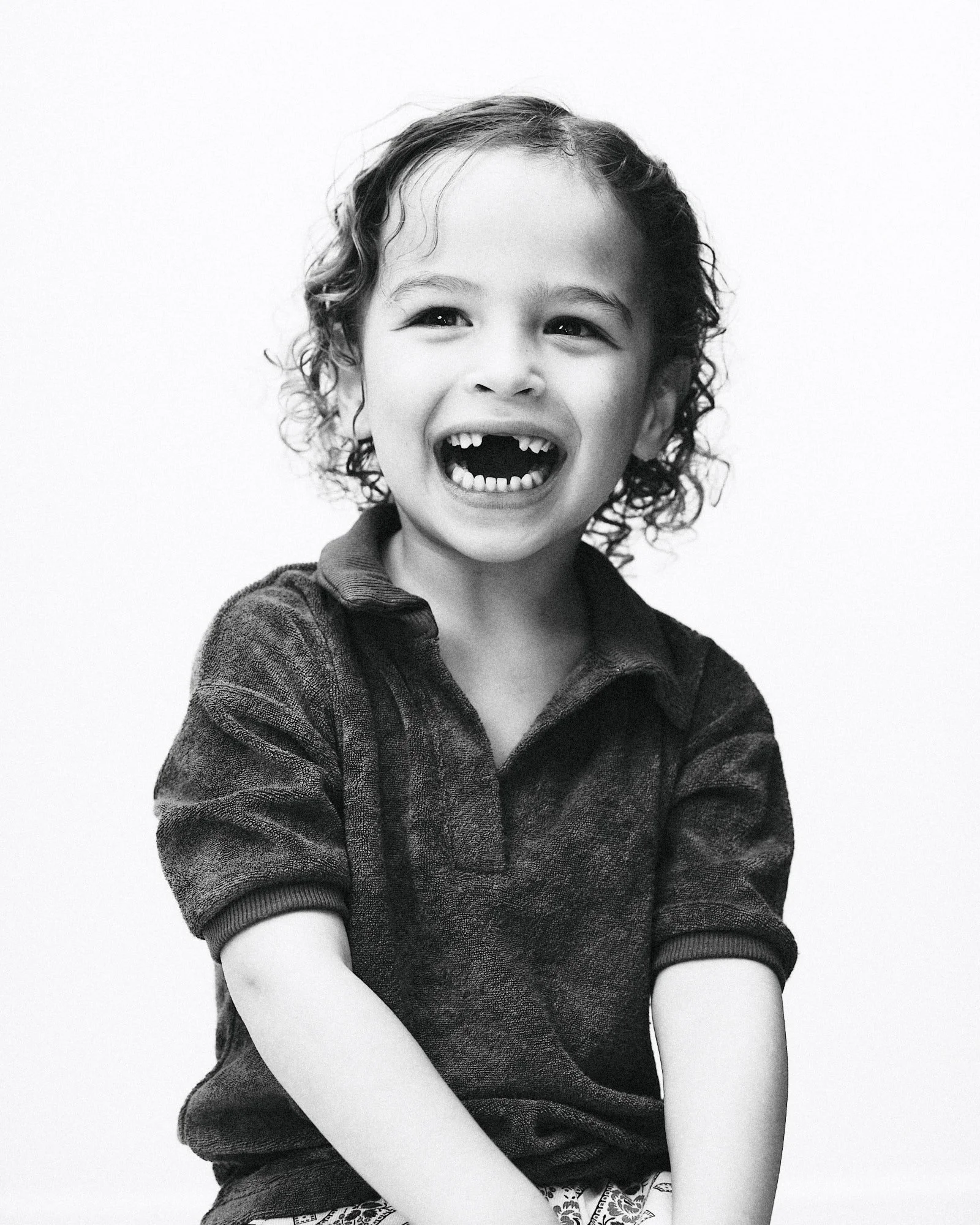 Black and white photo of a young girl smiling widely, showing her teeth, with curly hair, wearing a long-sleeved sweater and sitting against a plain background.
