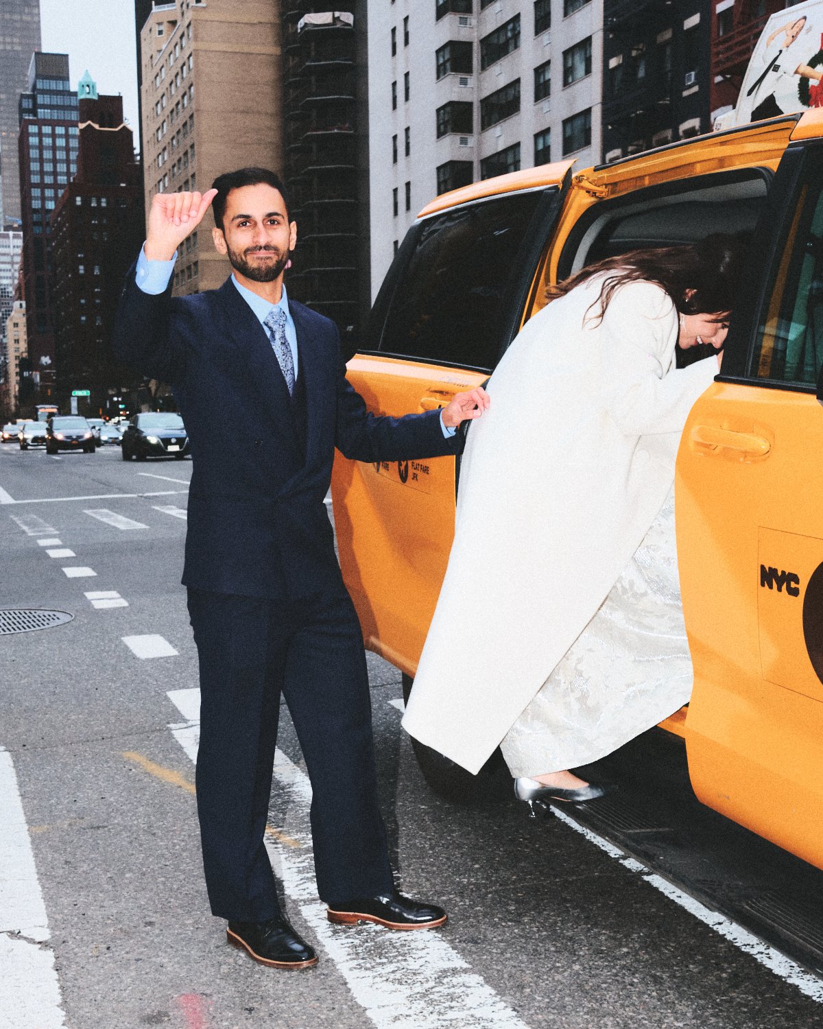 A man in a suit and a woman in a wedding dress standing outside near a yellow NYC taxi cab photographed by Madivan Studios, with the woman leaning into the backseat.