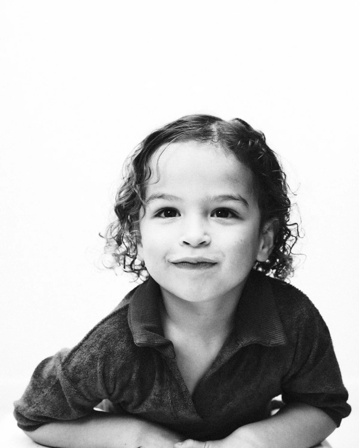 Black and white photo of a young girl with curly hair, smiling, wearing a dark shirt with a collar, against a plain background. Photographed in monochrome by Madivan Studios in New York City.