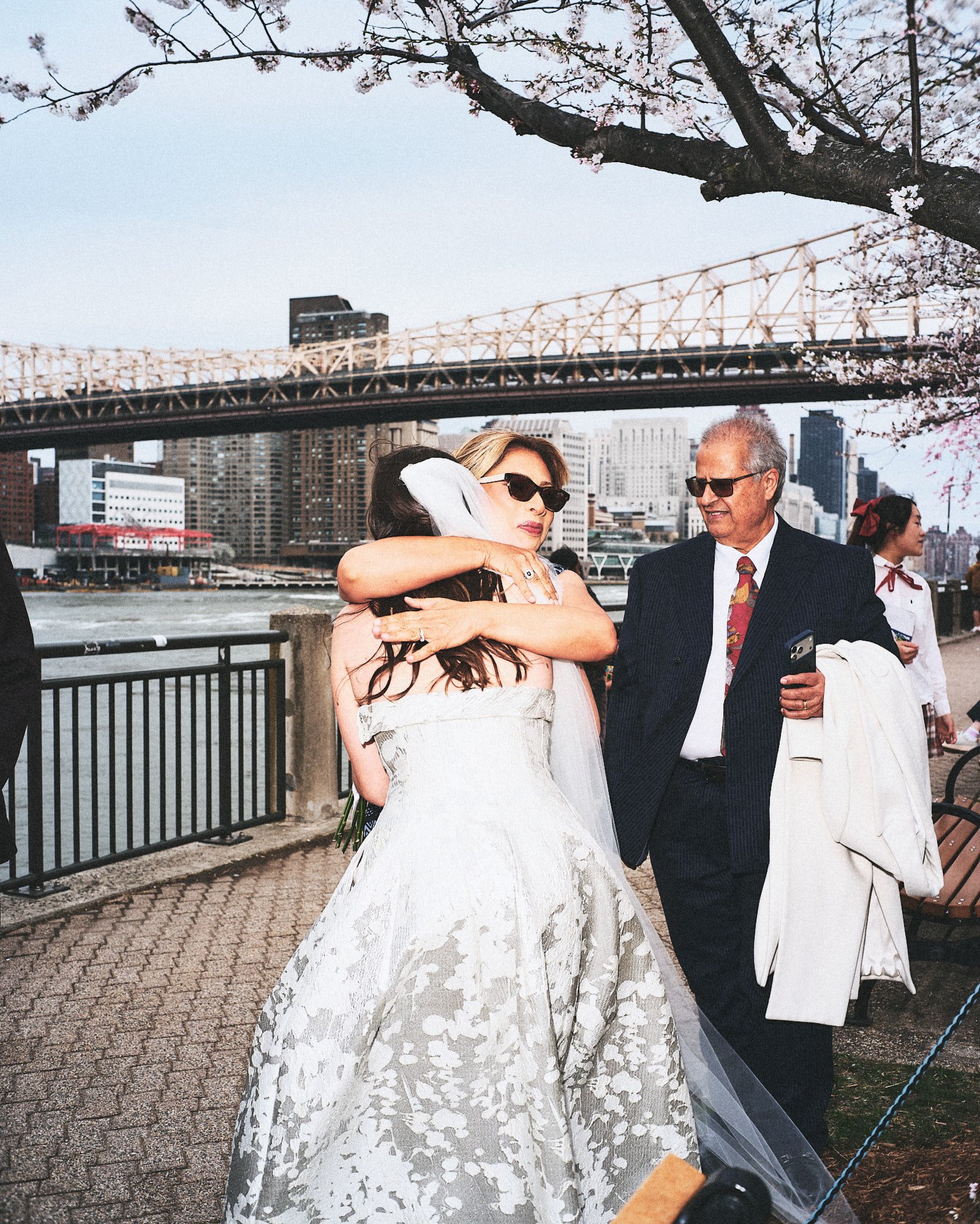 A woman in a white lace wedding dress embracing an older man in a dark suit and sunglasses by a riverside, with a city skyline, a bridge, and cherry blossom trees in the background.