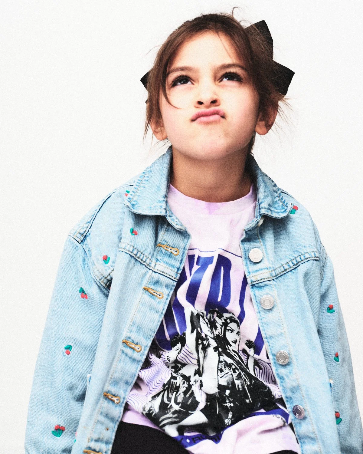 A young girl with brown hair and a black bow, making a pouting expression, wearing a denim jacket with small embroidered cherries, and a graphic T-shirt. Photographed in monochrome by Madivan Studios in New York City.