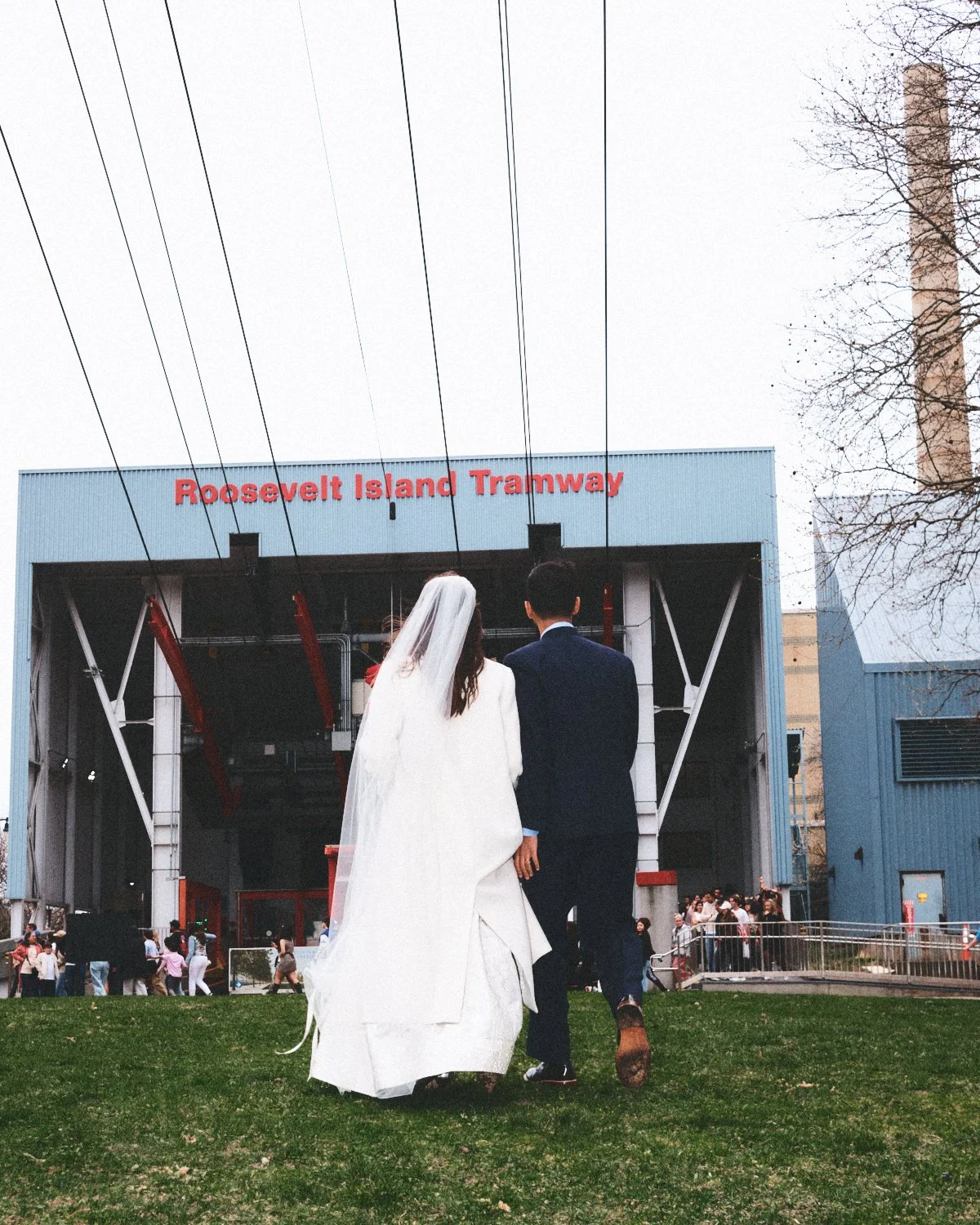 A bride and groom walking toward the Roosevelt Island Tramway station in New York City, with the station's sign visible, as people gather outside at their wedding. Photographed by Madivan Studios.