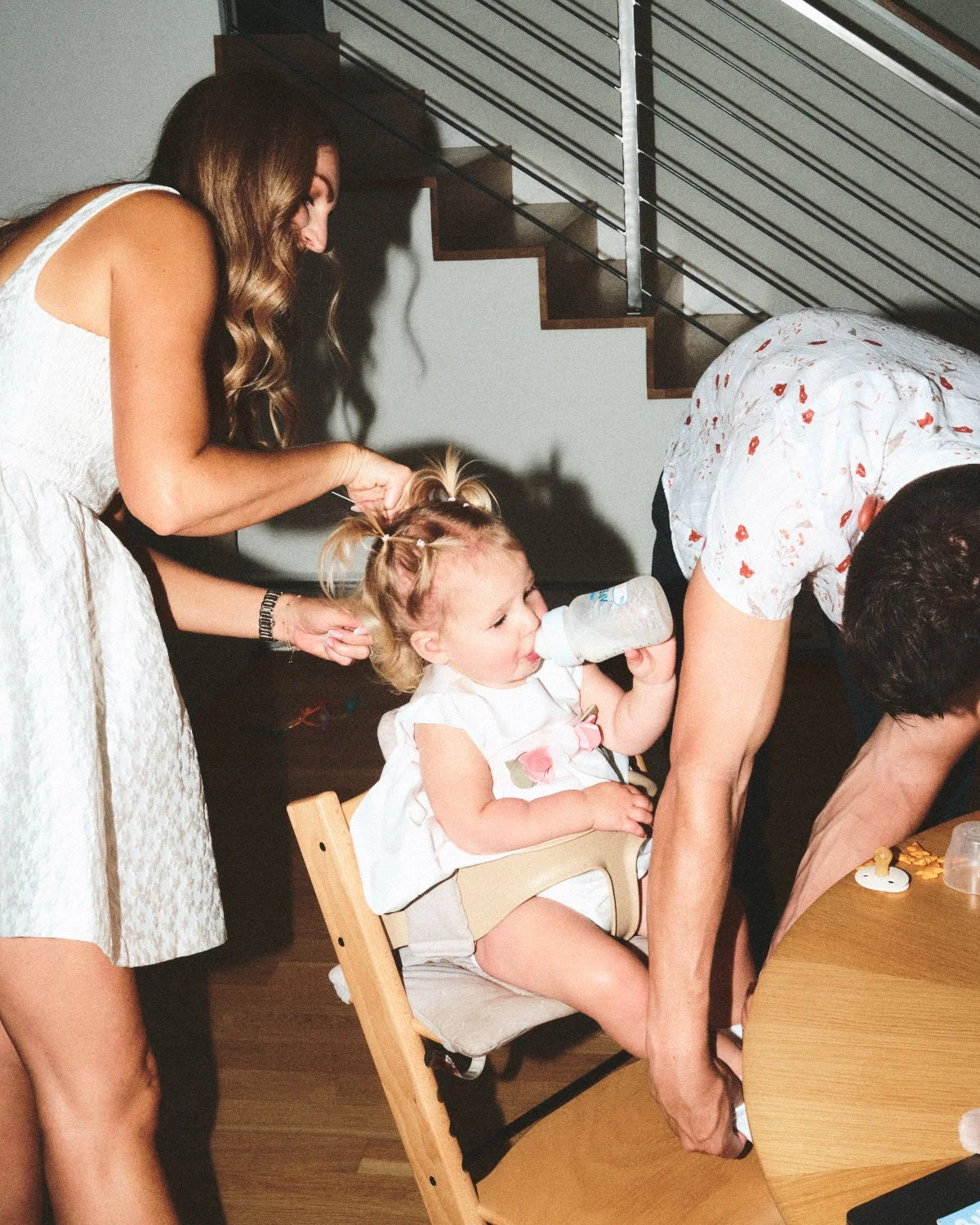 Documentary flash photography of a woman with long hair in a white dress is braiding a young girl’s blonde hair who is sitting in a high chair,  while a man bends over nearby. The scene is in a NYC apartment and photographed by Madivan Studios.