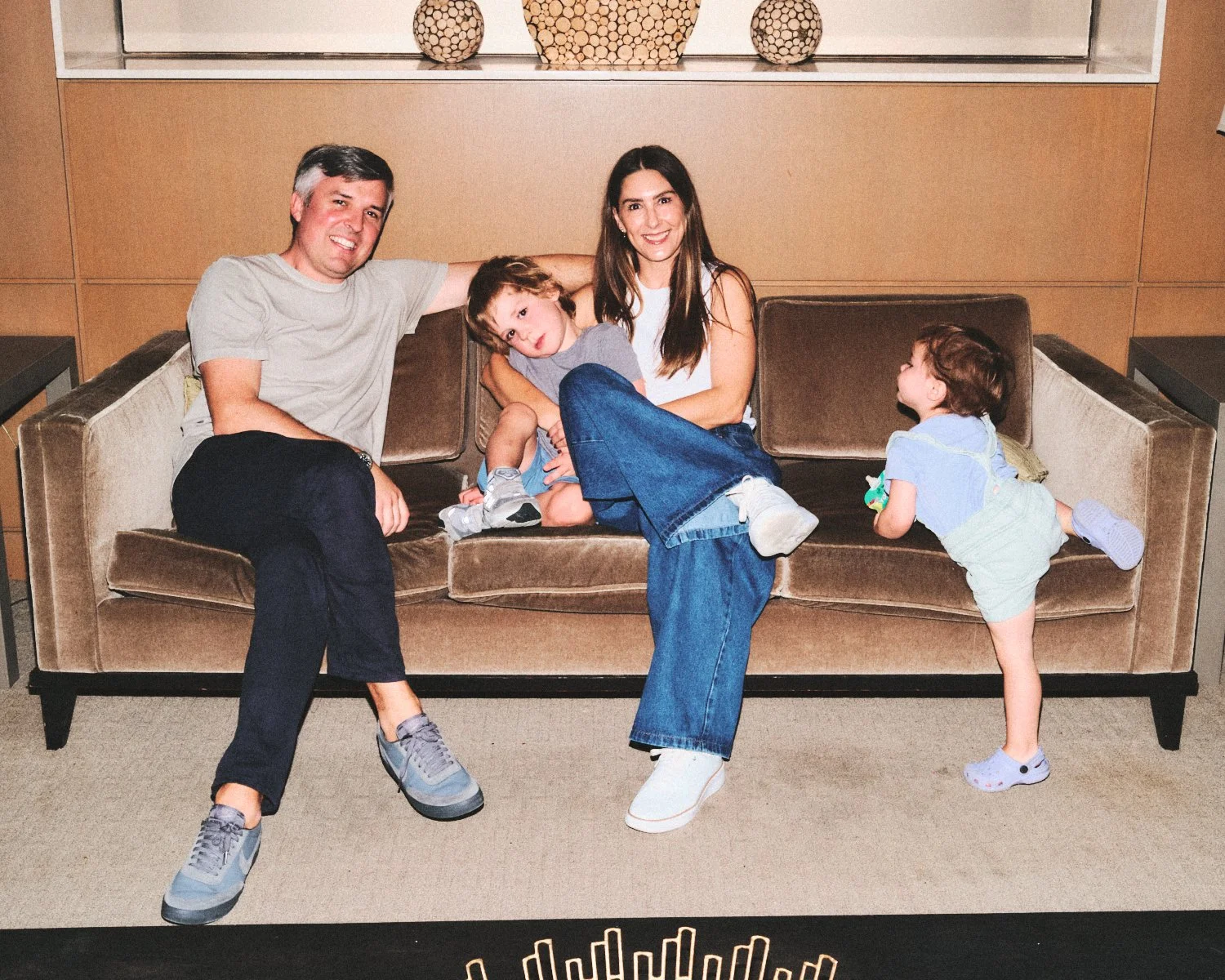 Documentary flash photography of a family sitting on a beige couch in a NYC living room. The father is smiling and wearing a gray t-shirt and dark pants. The mother is also smiling, they are being photographed by Madivan Studios.