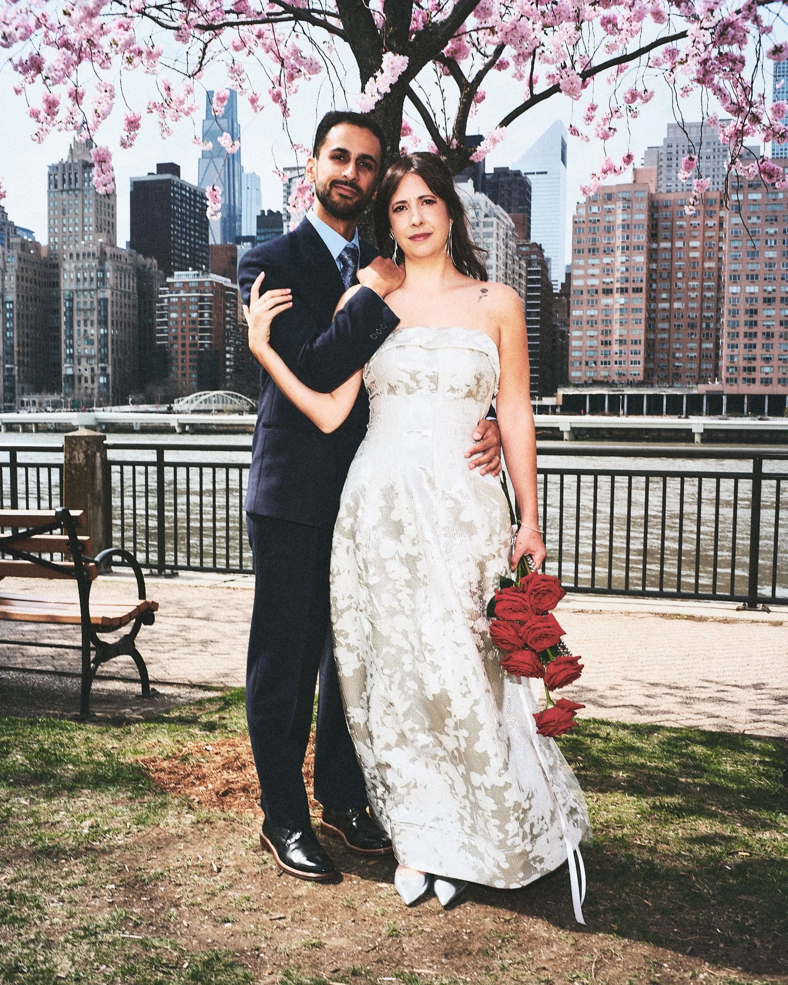 A couple dressed in formal wedding attire standing outdoors under a pink flowering tree with city buildings in the background. The woman is holding a bouquet of red roses and wearing a white strapless wedding gown, while the man is in a navy blue suit and tie.