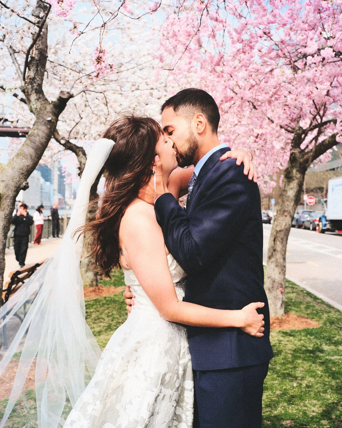 A bride and groom kissing under blooming cherry blossom trees during their wedding ceremony in a New York City outdoor park. Photographed by Madivan Studios.