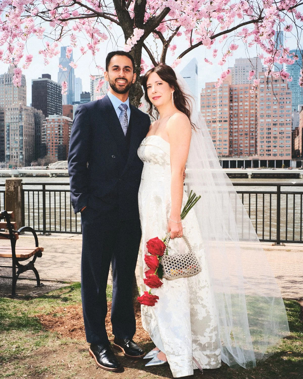 A bride and groom standing outdoors beside a river with NYC buildings in the background, near a cherry blossom tree. The bride is holding red roses and a small beaded handbag, wearing a white wedding dress and veil. Photographed by Madivan Studios.