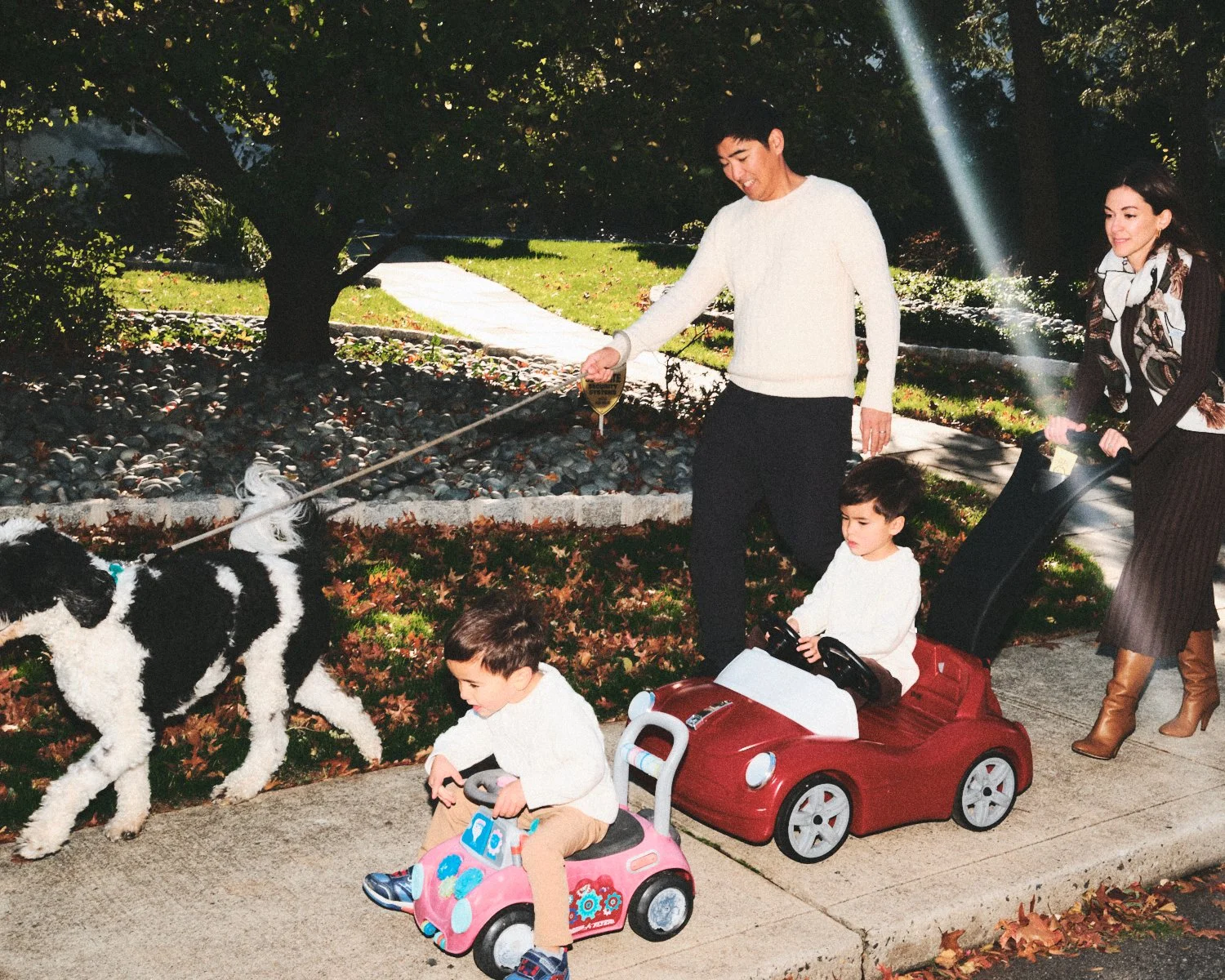 Documentary flash photography of a family walking in a NYC park with children riding toy cars and a dog on a leash. A Mom is walking behind them. The children are on small, ride-on toy cars, and they are being photographed by Madivan Studios.
