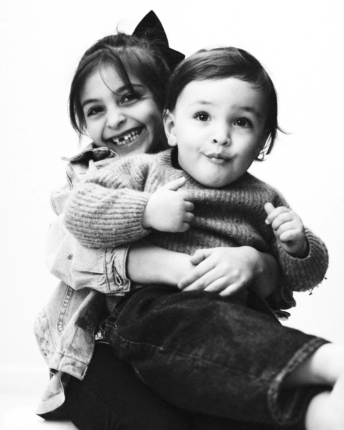 Black and white photo in a NYC photography studio of a young girl holding a young boy in her arms, both smiling and looking at the camera. Photographed in monochrome by Madivan Studios.