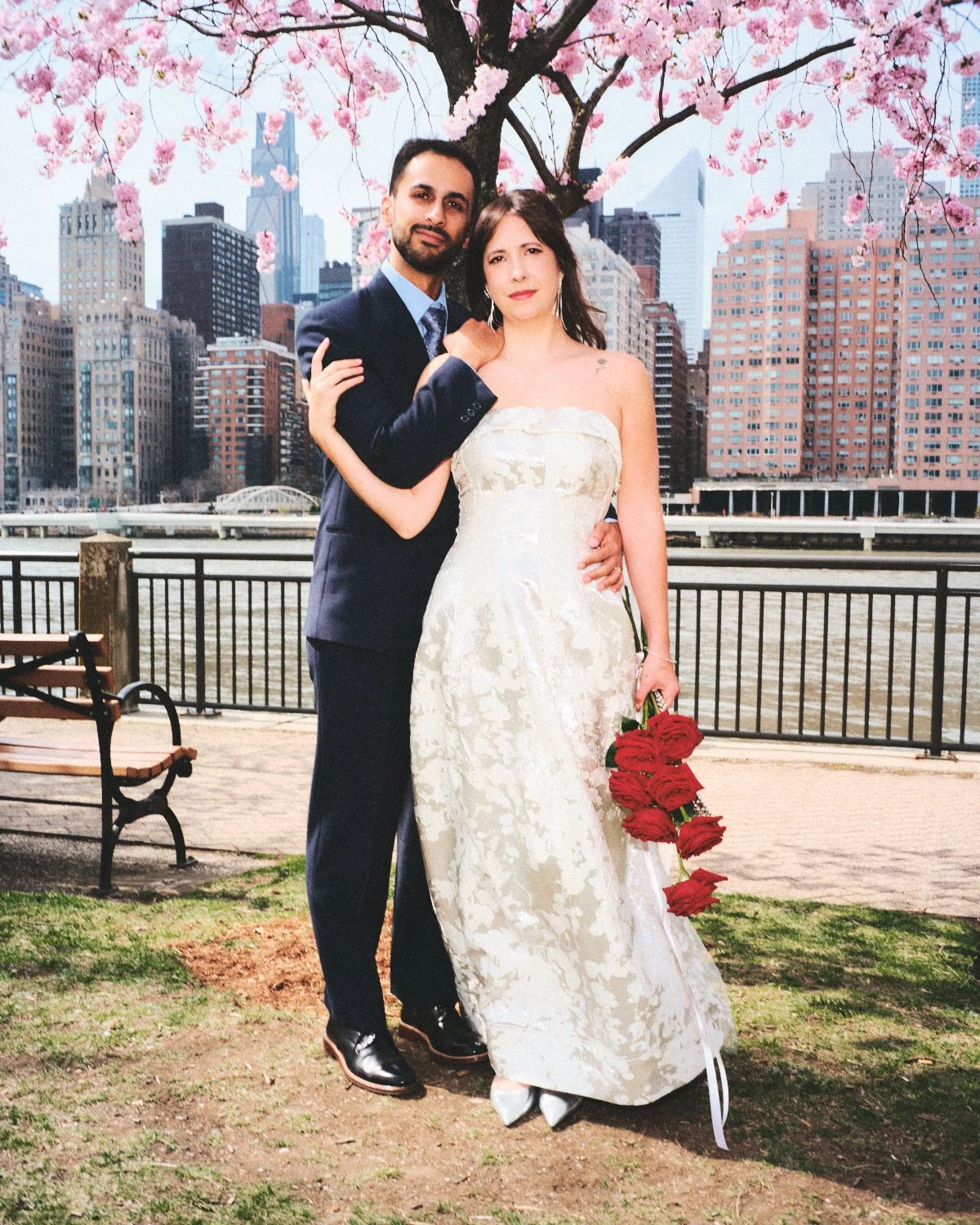 A newlywed couple under a blooming cherry blossom tree on a NYC riverfront promenade, with city skyscrapers in the background. The bride wears a strapless wedding gown and holds a bouquet of red roses, photographed by Madivan Studios.
