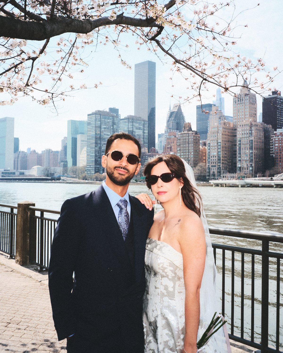 A couple dressed in wedding attire standing by the river with the NYC skyline in the background photographed by Madivan Studios, under a cherry blossom tree.
