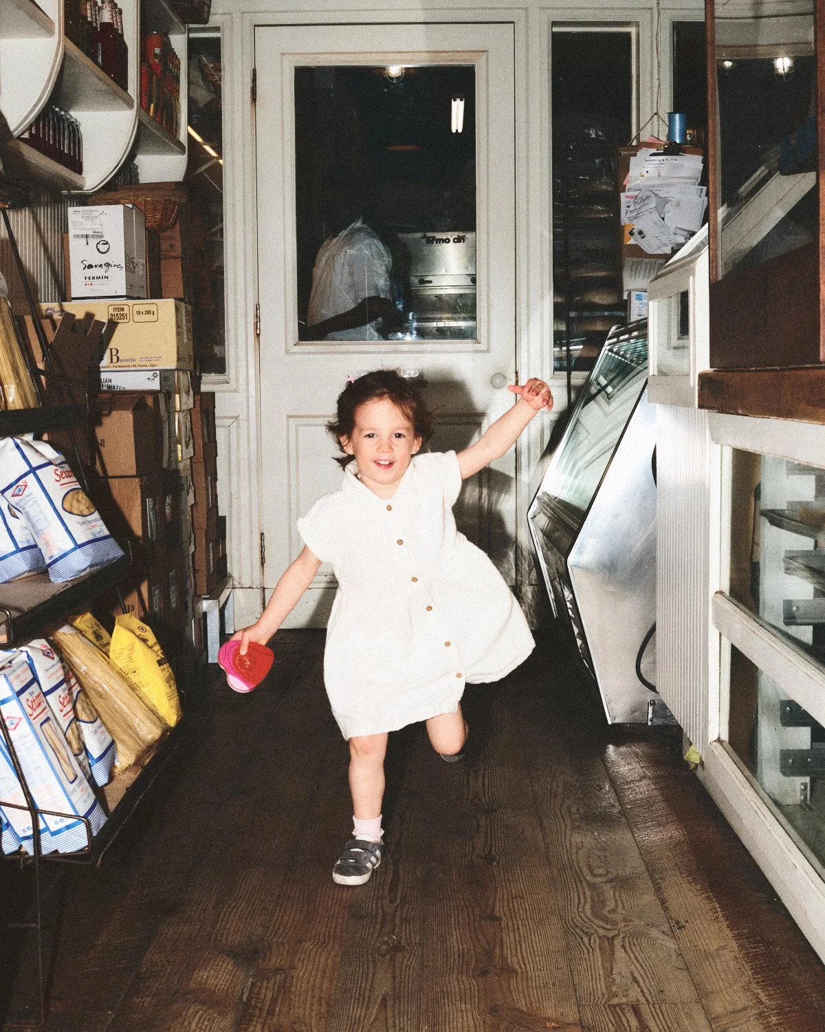 Documentary flash photography of a young girl with wearing a dress, and pink socks is happily dancing in a grocery store. The Saraghina store shelves are stocked, and she is being photographed by Madivan Studios.