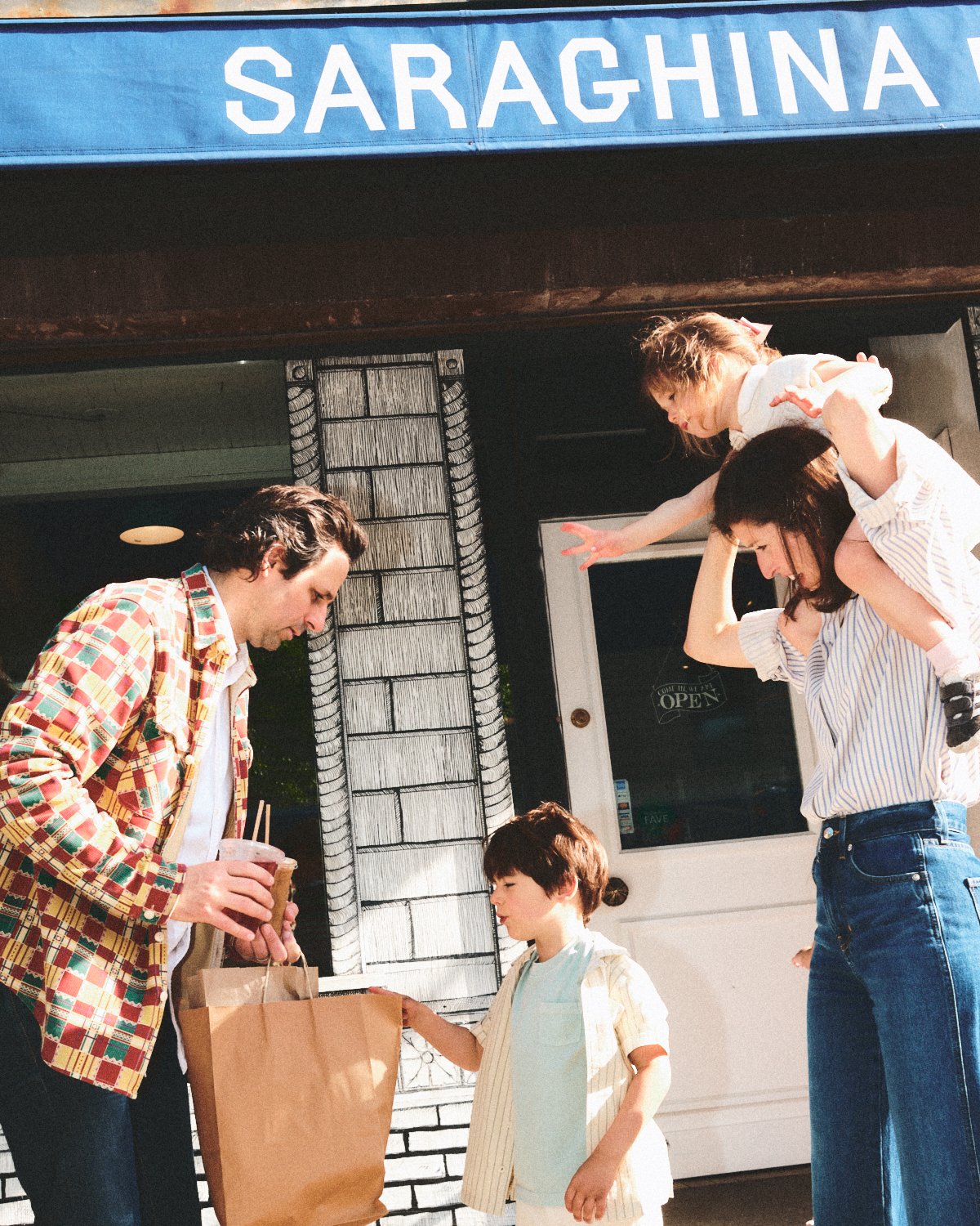 Documentary flash photography of a family enjoying outside in NYC at a shop with a sign reading 'SARAGHINA'. The father is handing a bag to a young boy, while the mother is giving her daughter a shoulder ride, photographed by Madivan Studios.