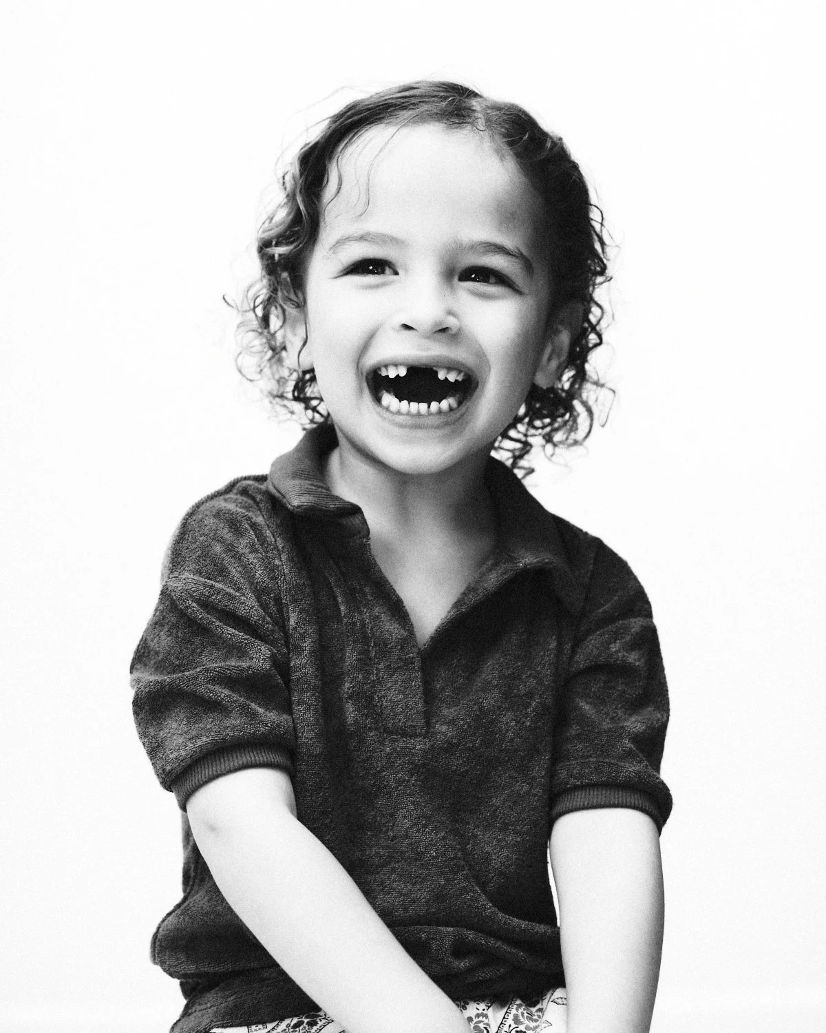 Black and white photo of a happy young boy with curly hair, smiling widely, showing missing teeth, wearing a sweatshirt. Photographed in monochrome in a NYC studio by Madivan Studios in New York City.