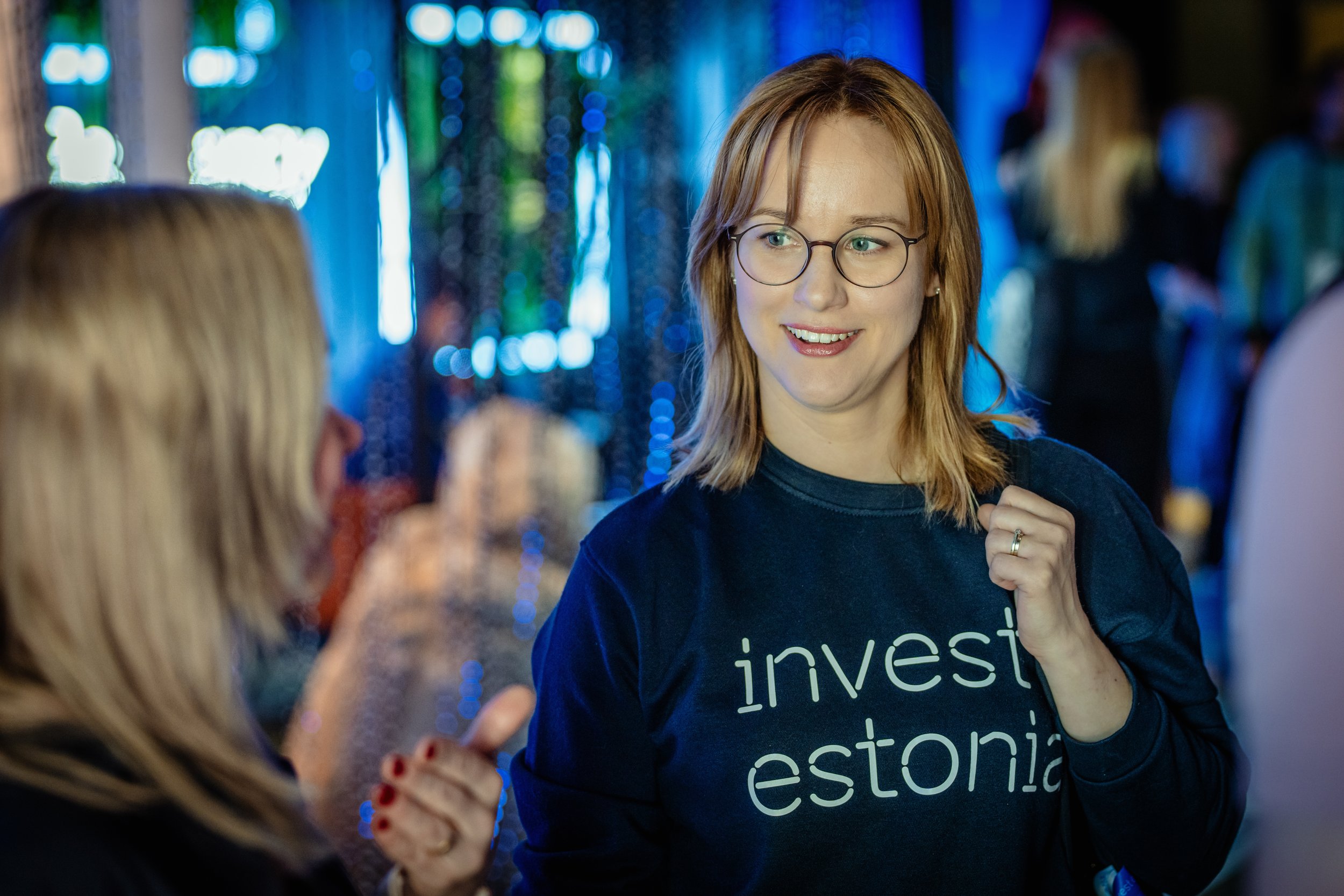 A young woman with red hair and glasses smiling and talking with another person at an indoor event, wearing a black shirt with the words 'invest estonia'.