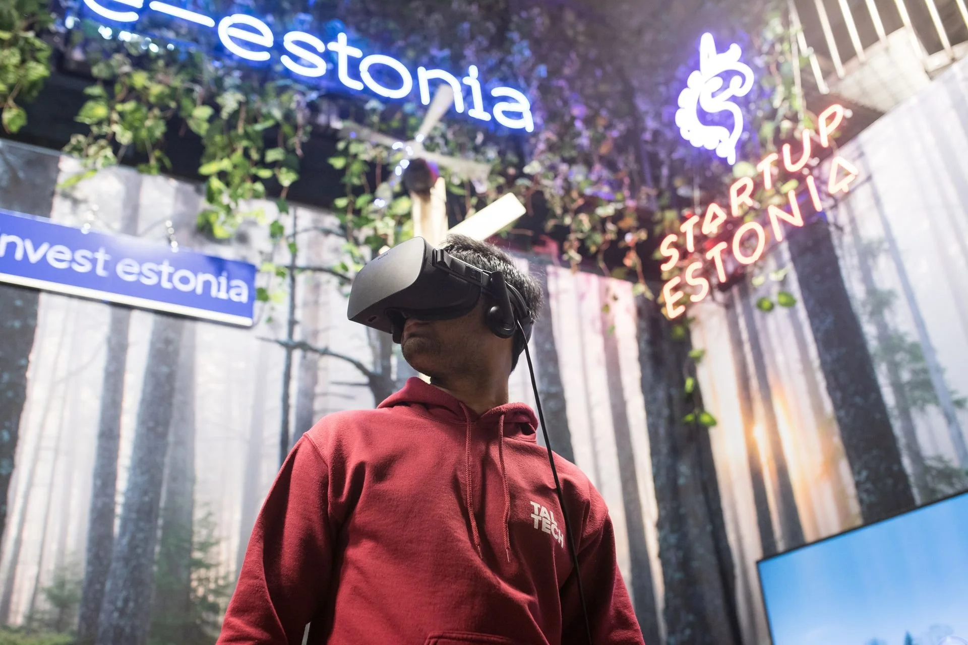 A man in a red hoodie with a headset and wearing a virtual reality headset in an indoor exhibit, with neon signs reading 'estonia' and 'startup estonia' and a forest backdrop.
