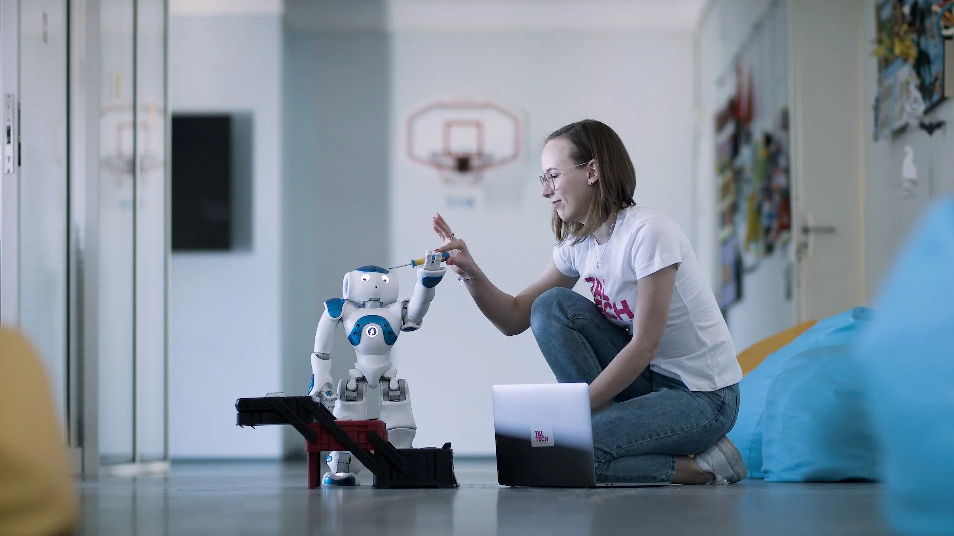 A young woman sitting on the floor in a school gymnasium, working on a robot with a screwdriver. She is smiling and wears glasses, a white T-shirt, and jeans. The robot is standing on a toolbox, with a laptop nearby.