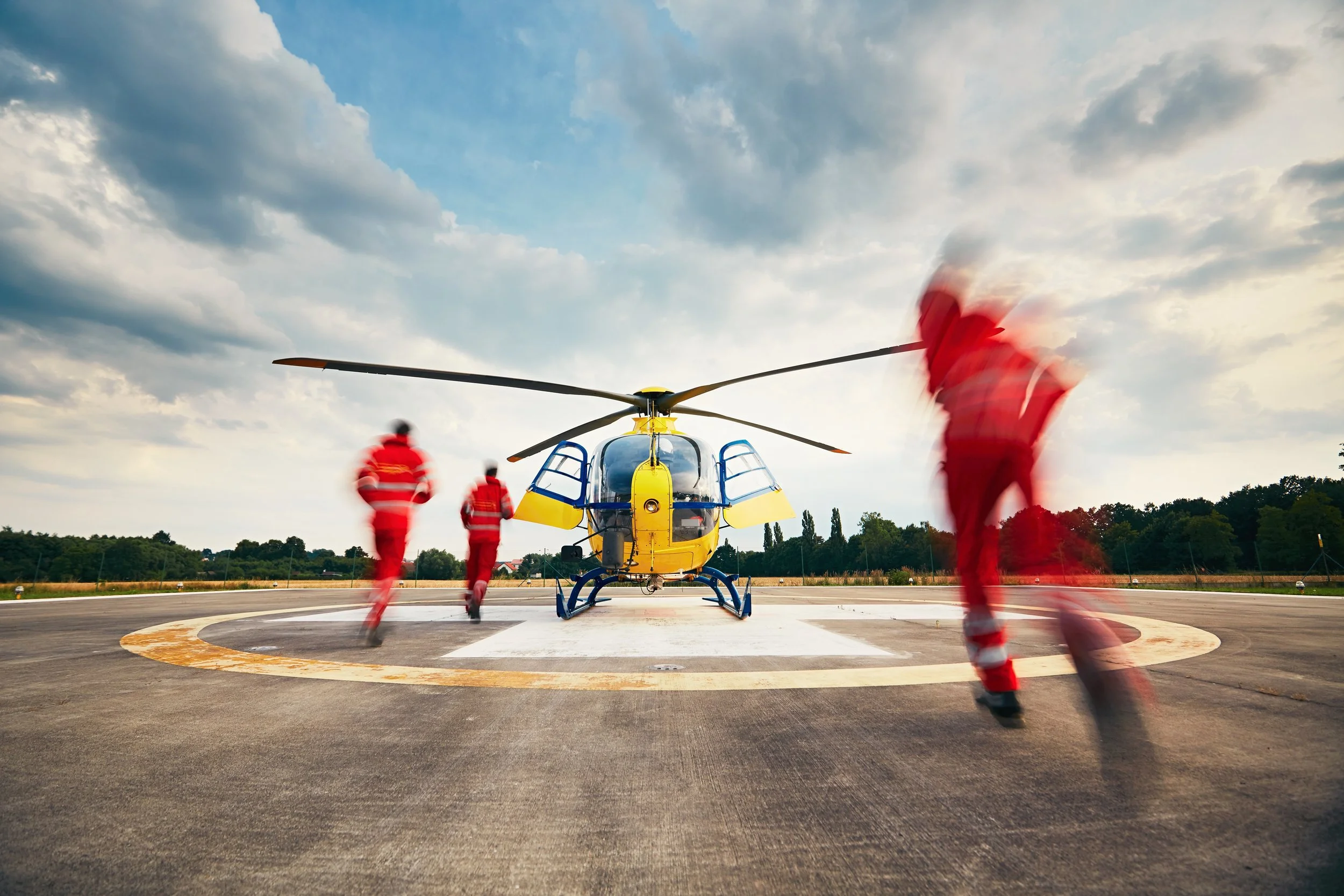 A yellow emergency helicopter on a helipad with three rescue workers in red uniforms walking towards it, under a partly cloudy sky.