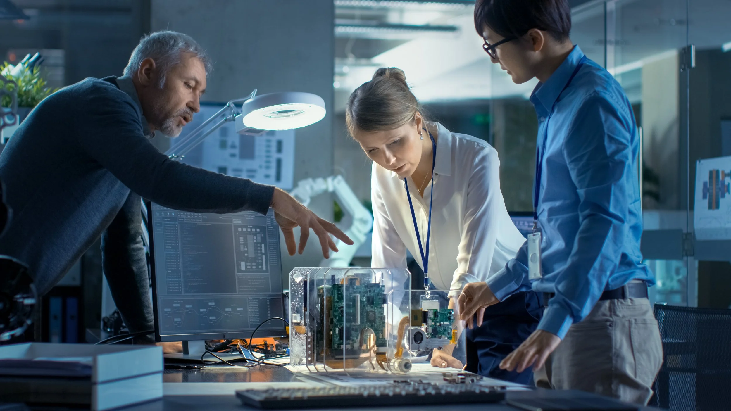 Three researchers working on electronic components in a laboratory, discussing a transparent enclosure containing circuit boards and wires.