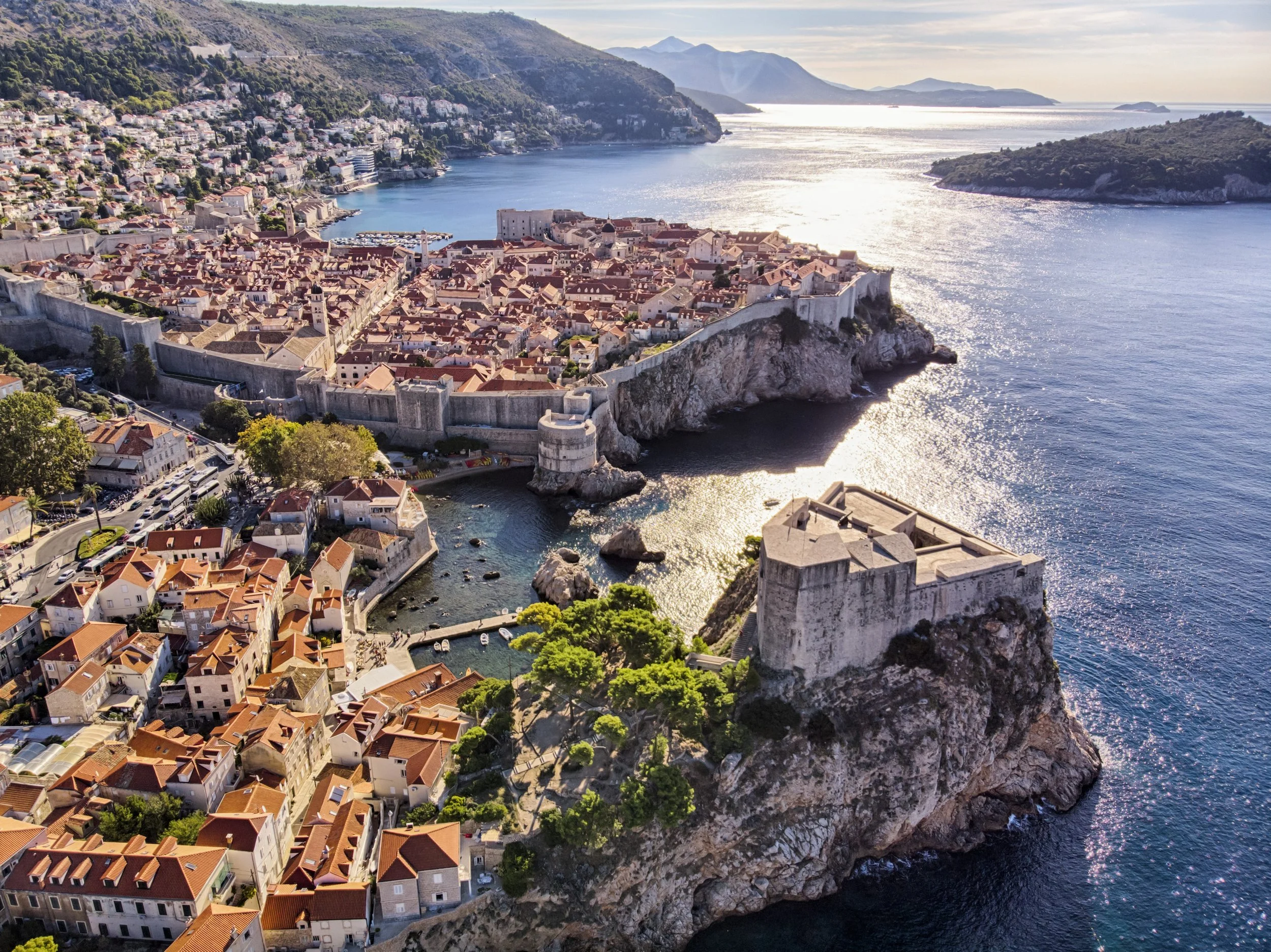 Aerial view of a historic coastal town with red-tiled roofs surrounded by a stone wall, overlooking the sea with mountains in the background, likely Dubrovnik, Croatia.