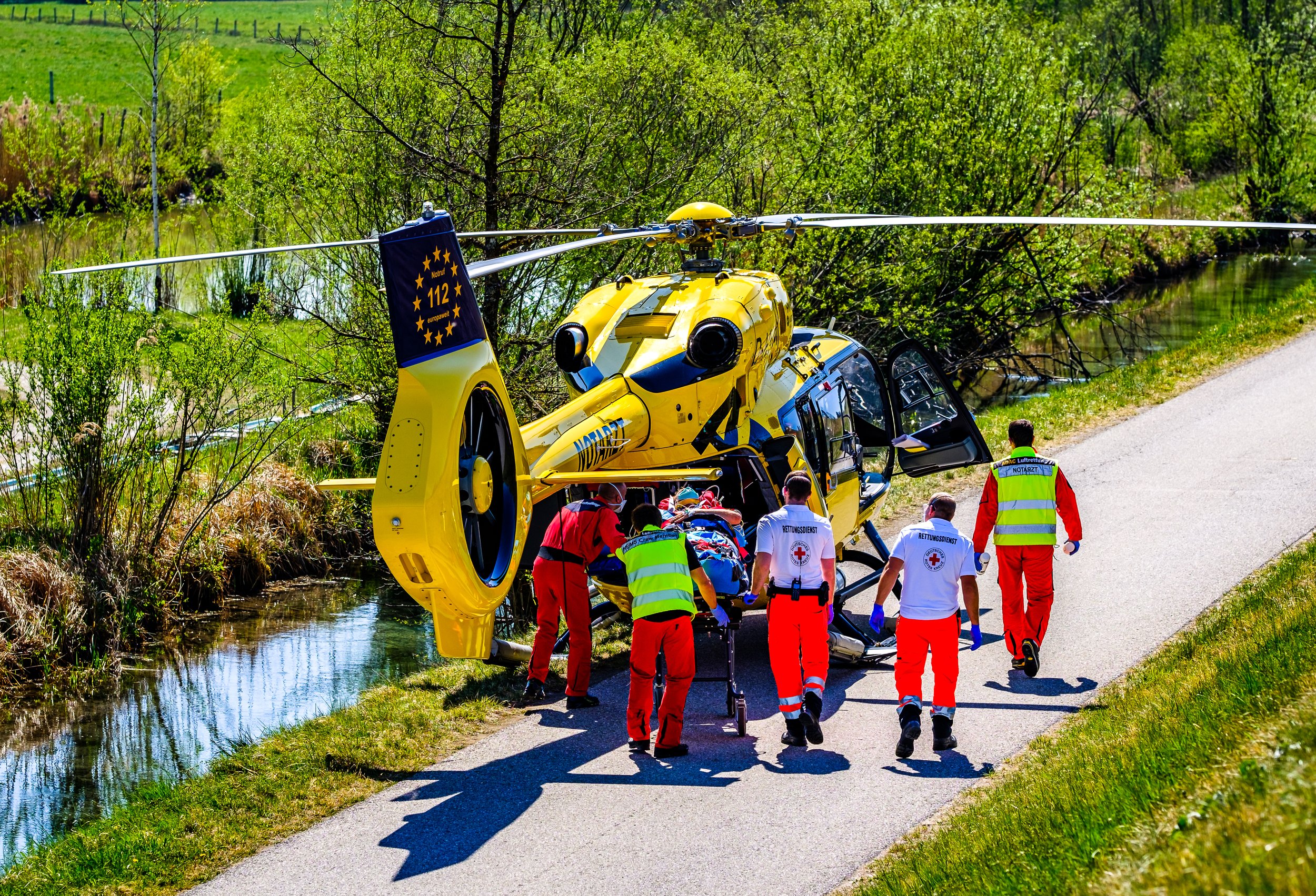 A yellow rescue helicopter is landed on a narrow road beside a small canal, with six medical personnel assisting a person on a stretcher.