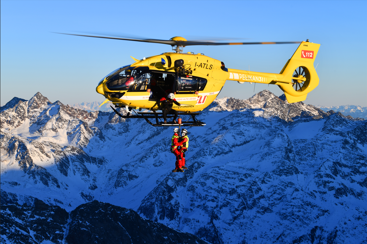 A yellow rescue helicopter flying over snow-covered mountains with two rescue workers hanging below.