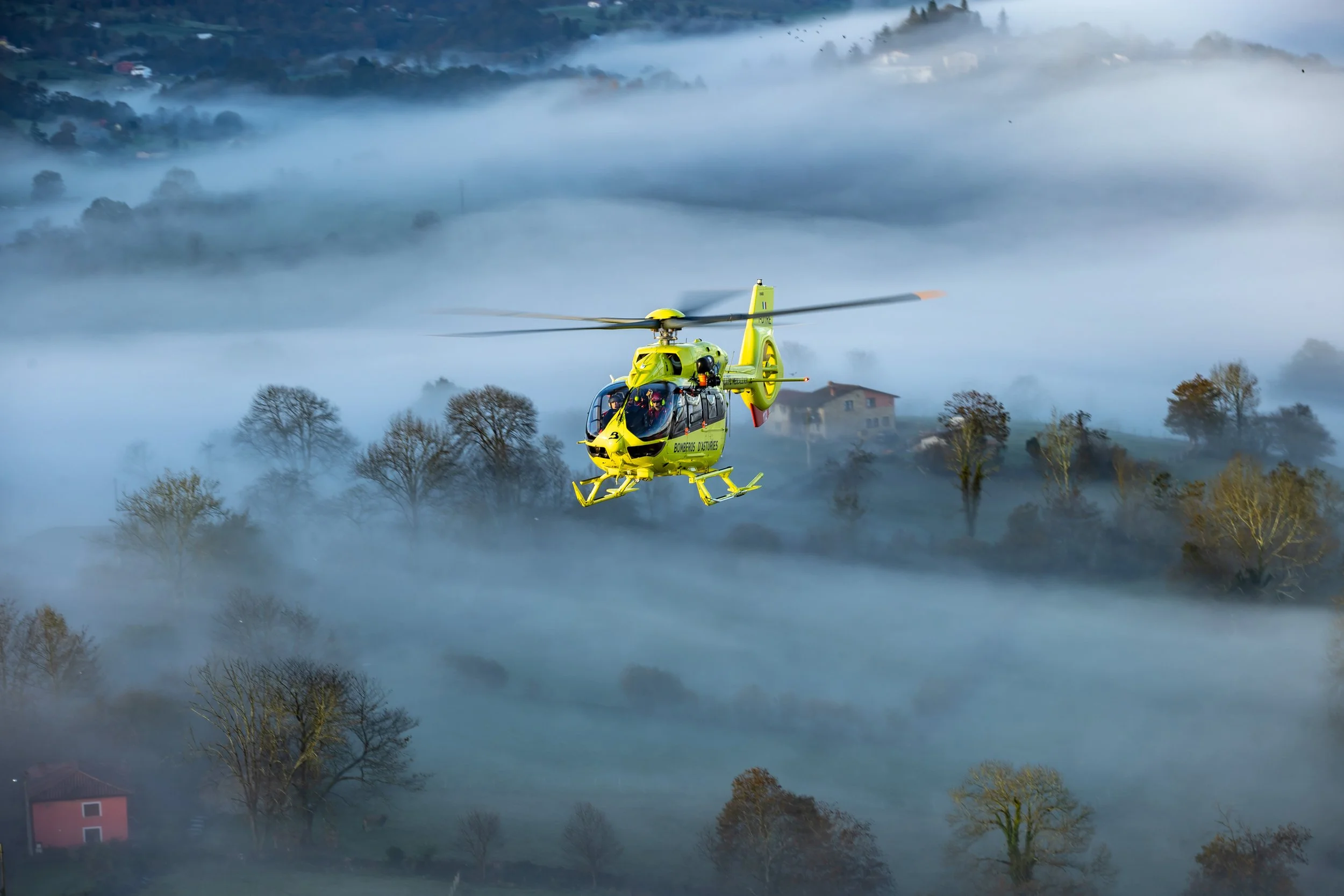 A yellow helicopter flying over a foggy landscape with trees and houses.