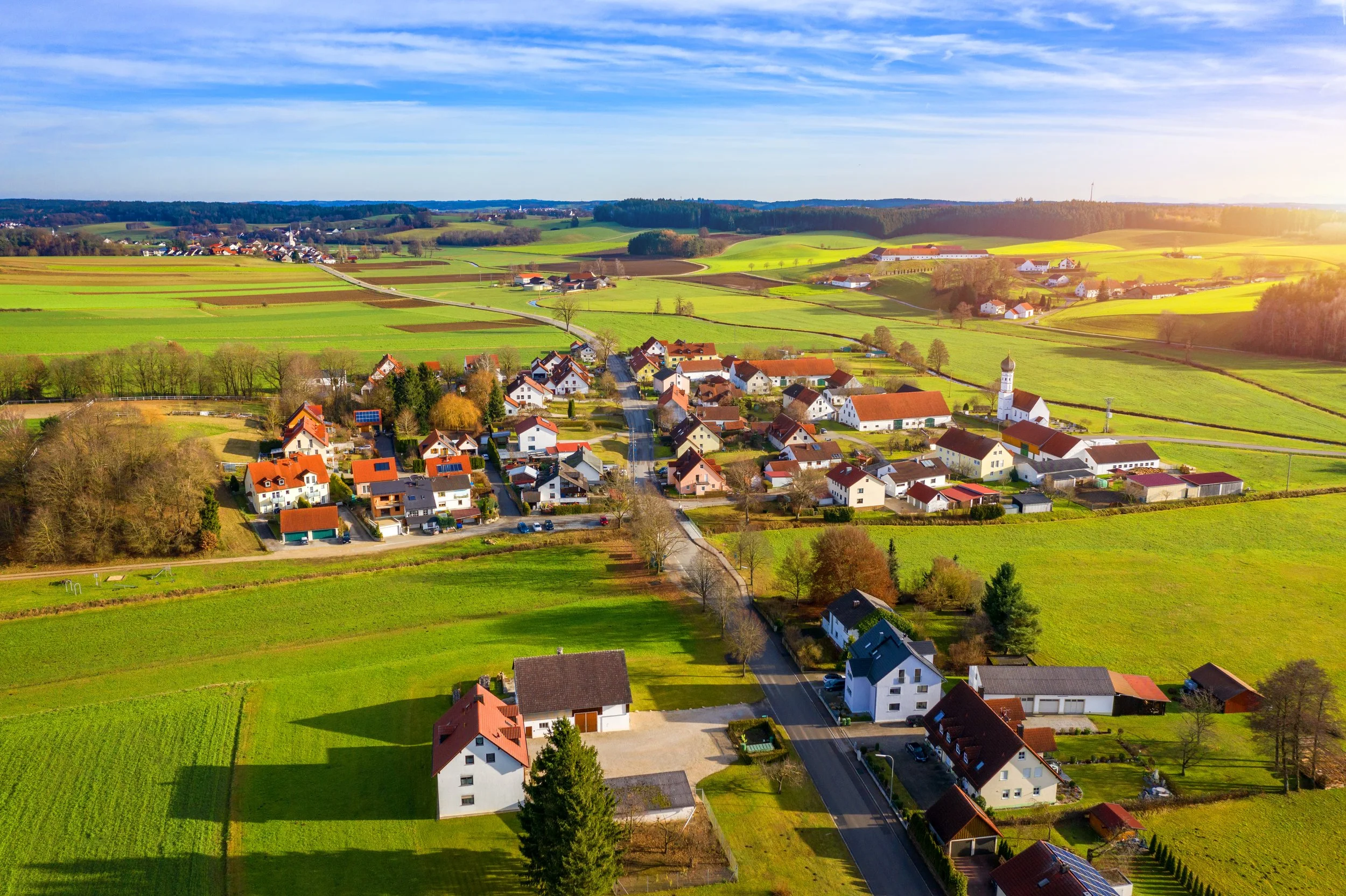 Aerial view of a small village surrounded by green fields and rolling hills, with a blue sky overhead.