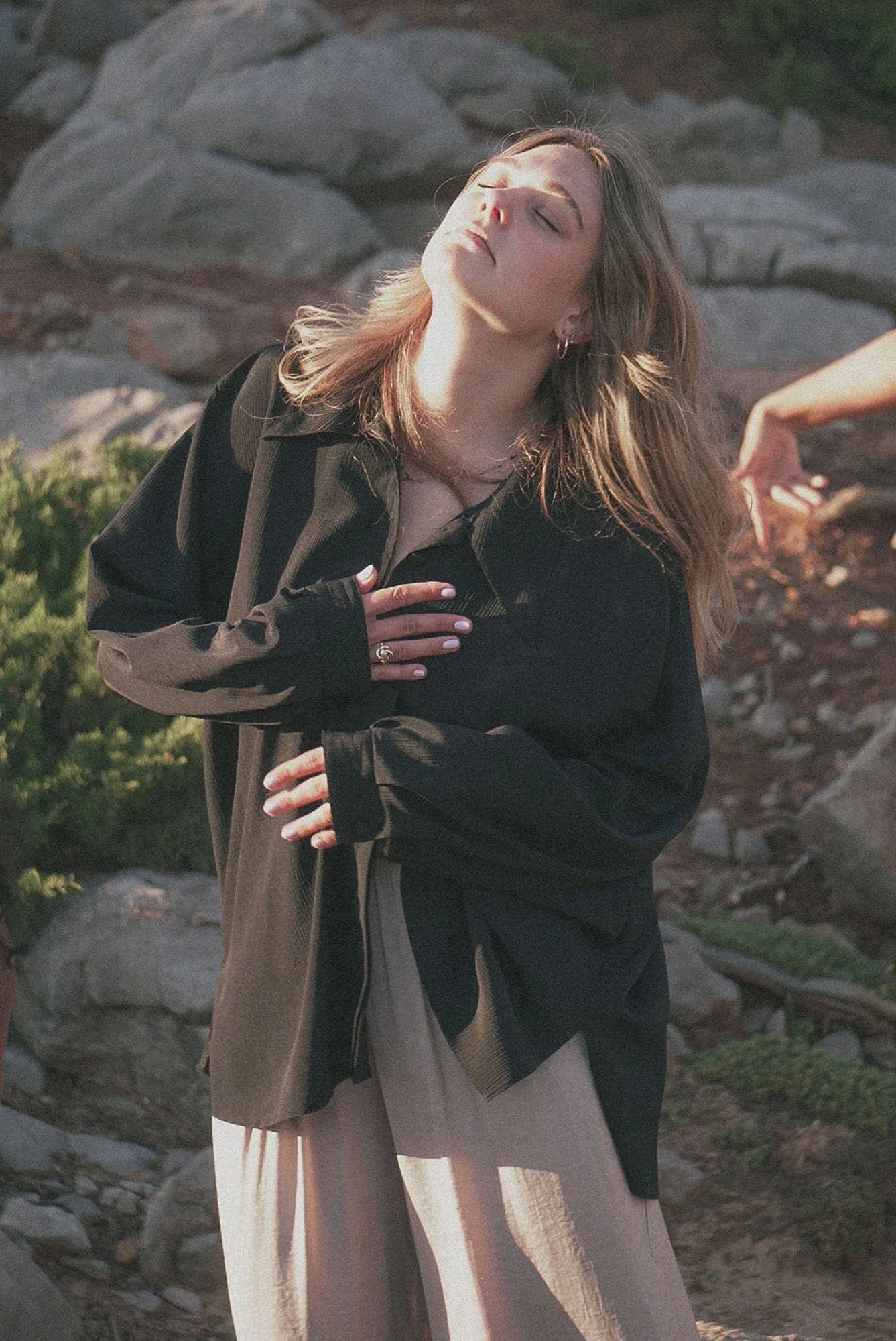 A woman, a breathwork facilitator a coach with light brown hair, wearing a black shirt and beige wide-leg pants, is outdoors with her eyes closed and one hand on her chest, surrounded by rocks and greenery.Wave breath, circular breath.