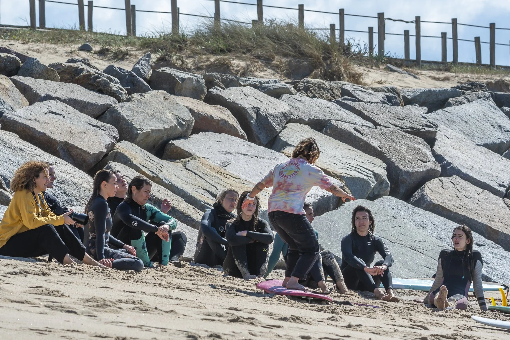 Group of people sitting on a beach in wetsuits, receiving a surfing tutorial from an instructor pointing to a surfboard. Porto Surf Femmes,  photography by Abilio Meneses
