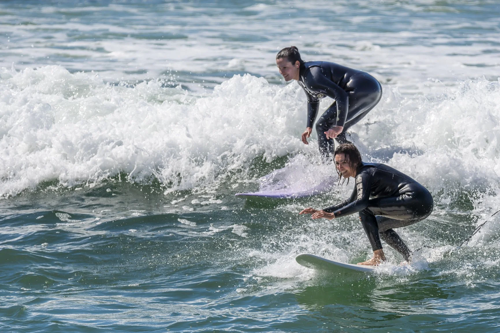 Two people surfing on ocean waves, wearing black wetsuits. Porto Surf Femmes,  photography by Abilio Meneses