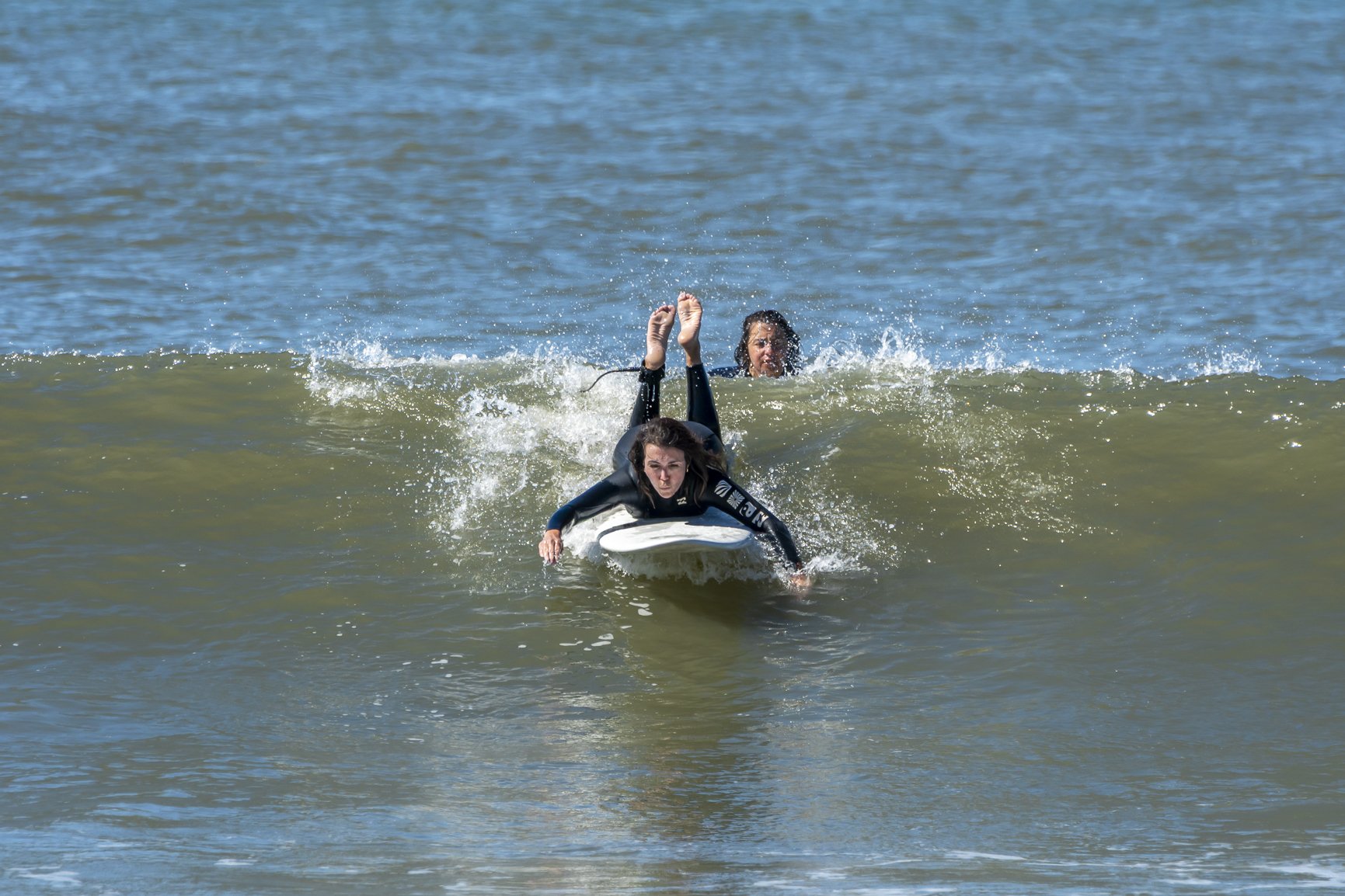 Two women wearing wetsuits surfing on a wave, one lying on a surfboard and the other behind, in the ocean. Porto Surf Femmes. Photography by Abilio Meneses