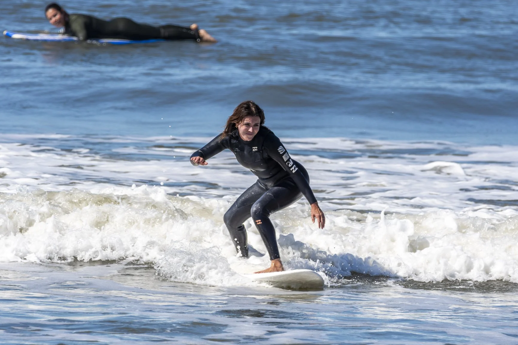 A woman surfing a small wave in the ocean, wearing a wetsuit, with another person on a surfboard in the background. Porto Surf Femmes. Photography by Abilio Meneses